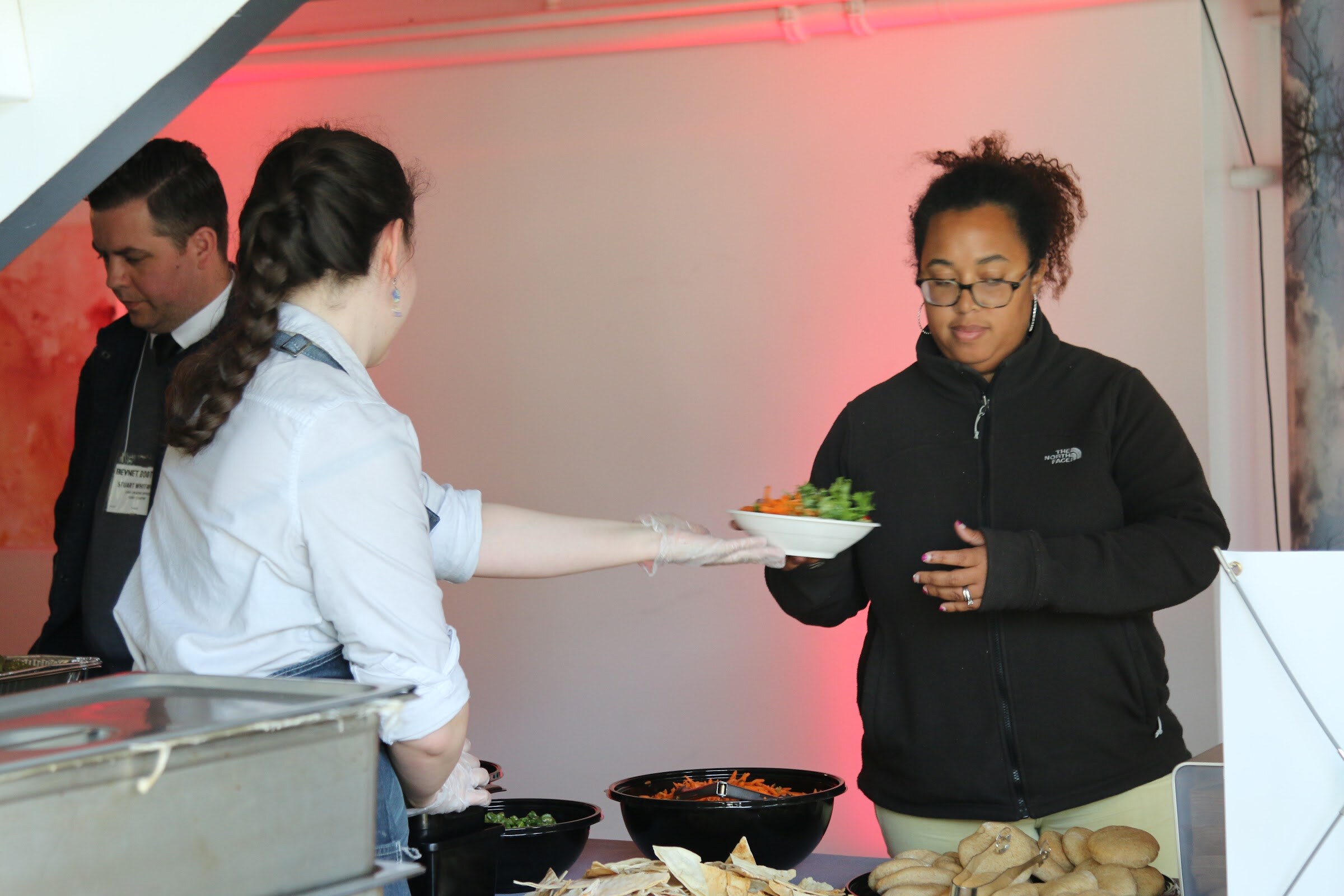 Server hands salad bowl to attendee at buffet under pink light.