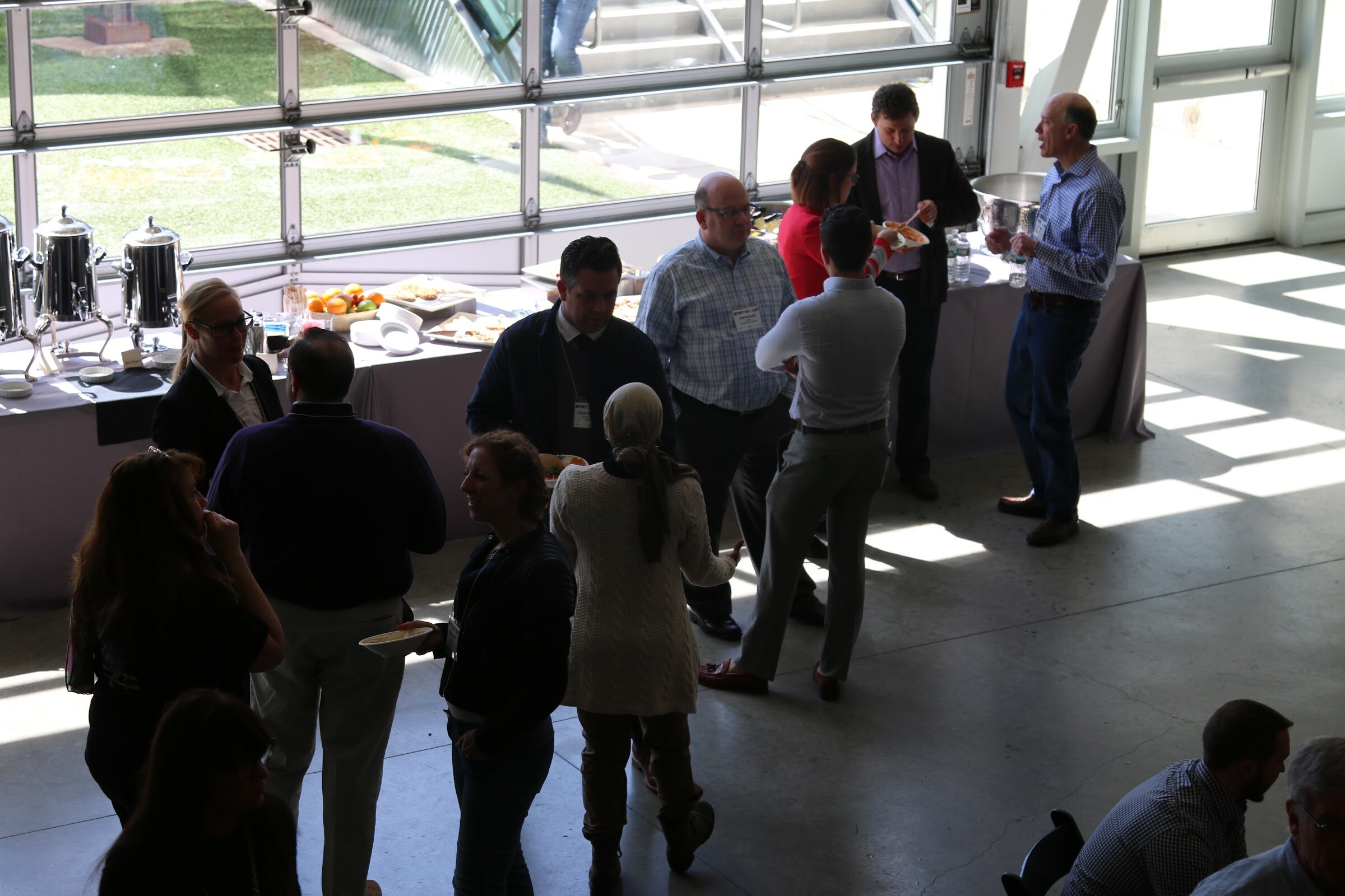 Elevated view of attendees lining up at lunch buffet beside coffee urns, sunlight through glass doors.