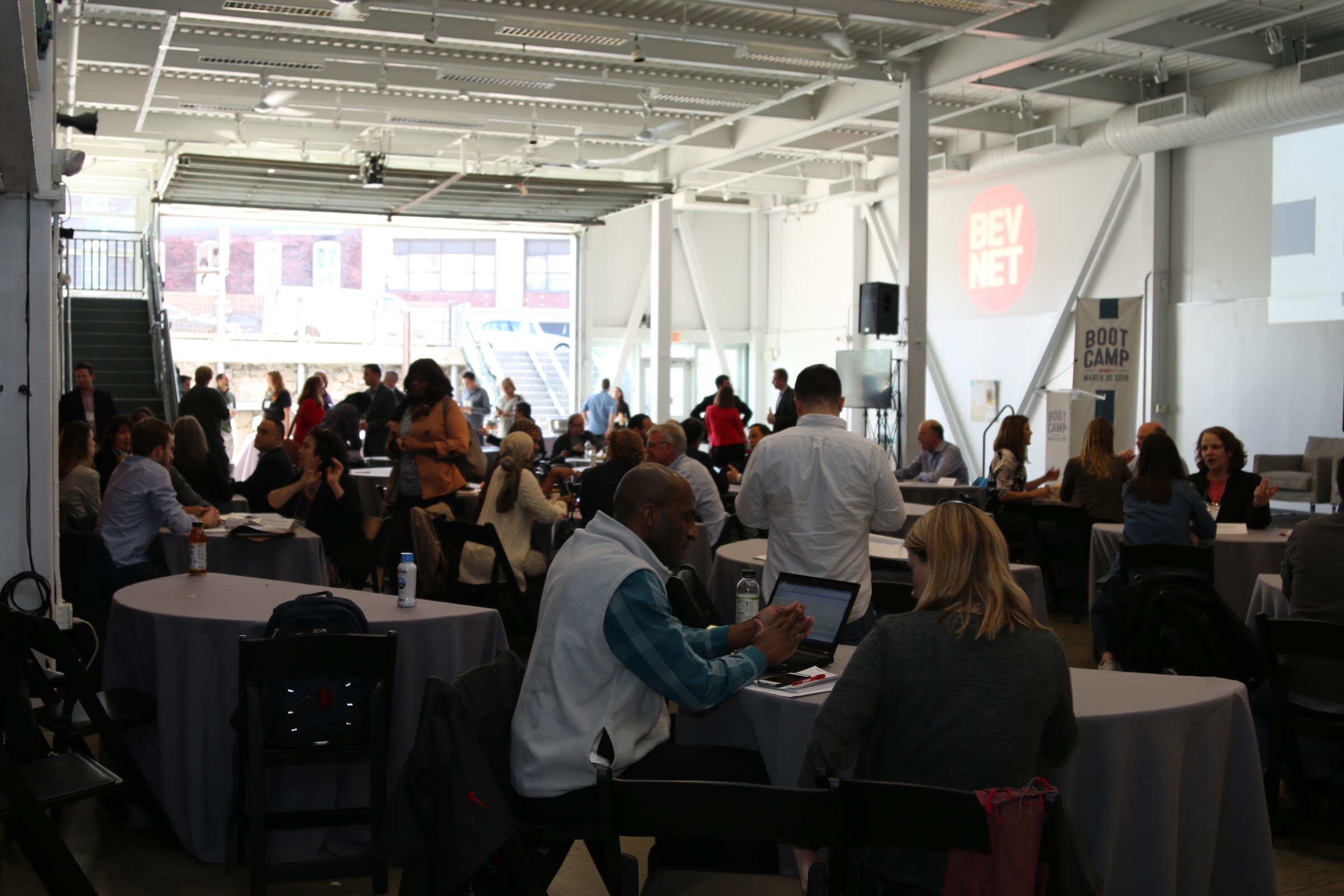 Wide room view of attendees in meetings and conversation, BevNET logo and Boot Camp banner at back.