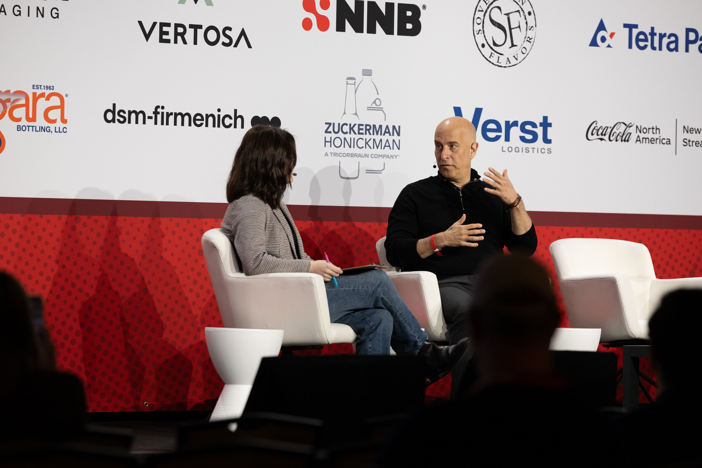 A woman and a man sit in white chairs on stage in front of a sponsor-logo backdrop.