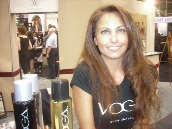 A smiling woman with long brown hair wears a black Voga Italia shirt beside several Voga bottles on a table, with other exhibitors visible in the background.