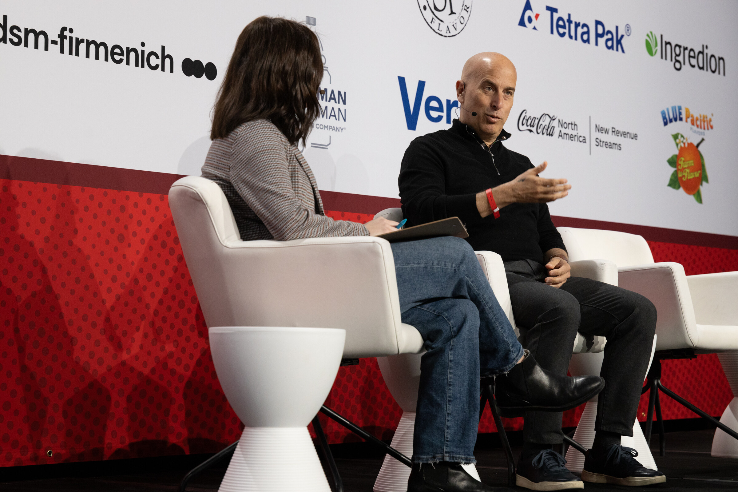 A woman and a man sit in white chairs on stage in conversation.