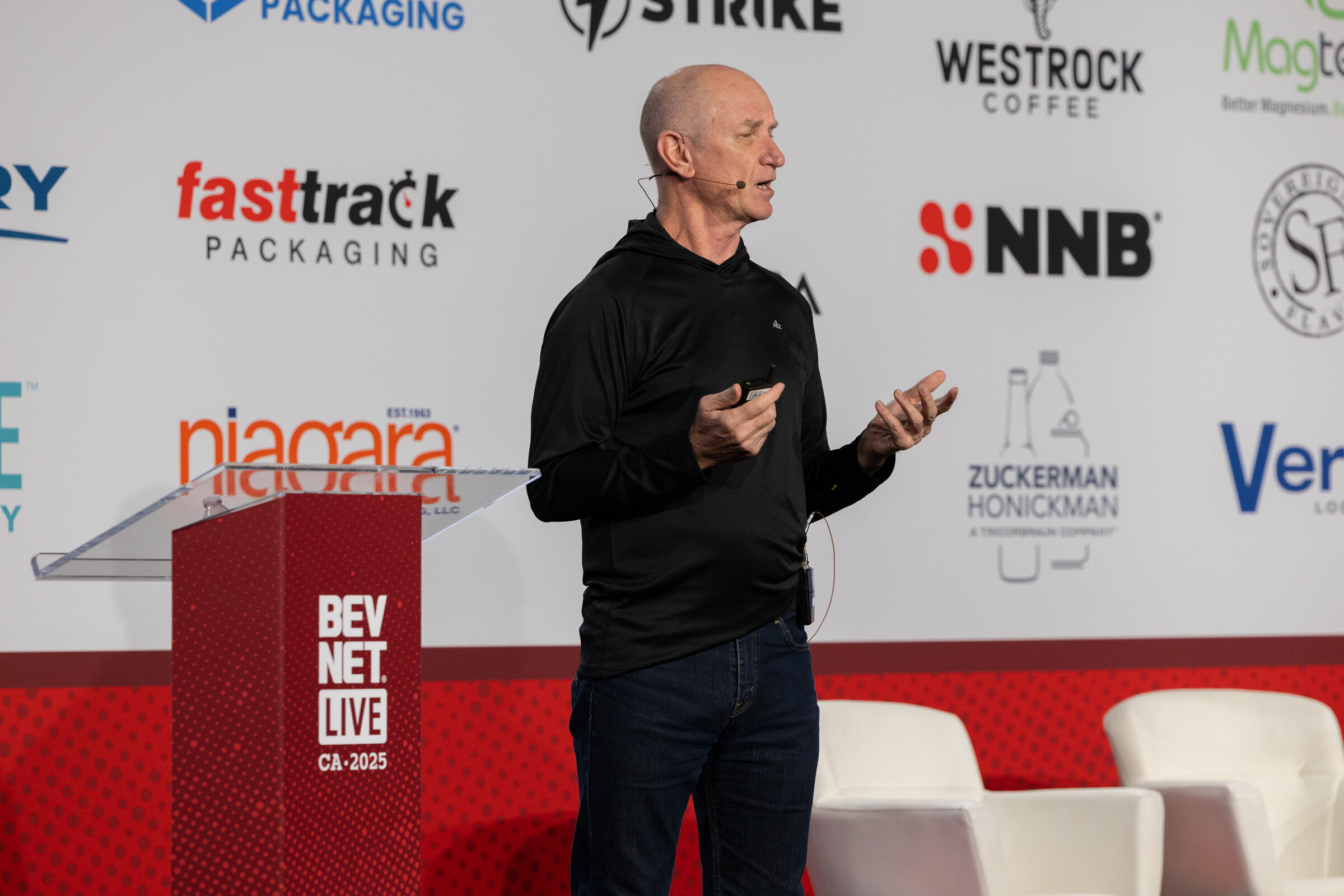 A man in a black shirt speaks and gestures beside a lectern on the Beverage School stage.