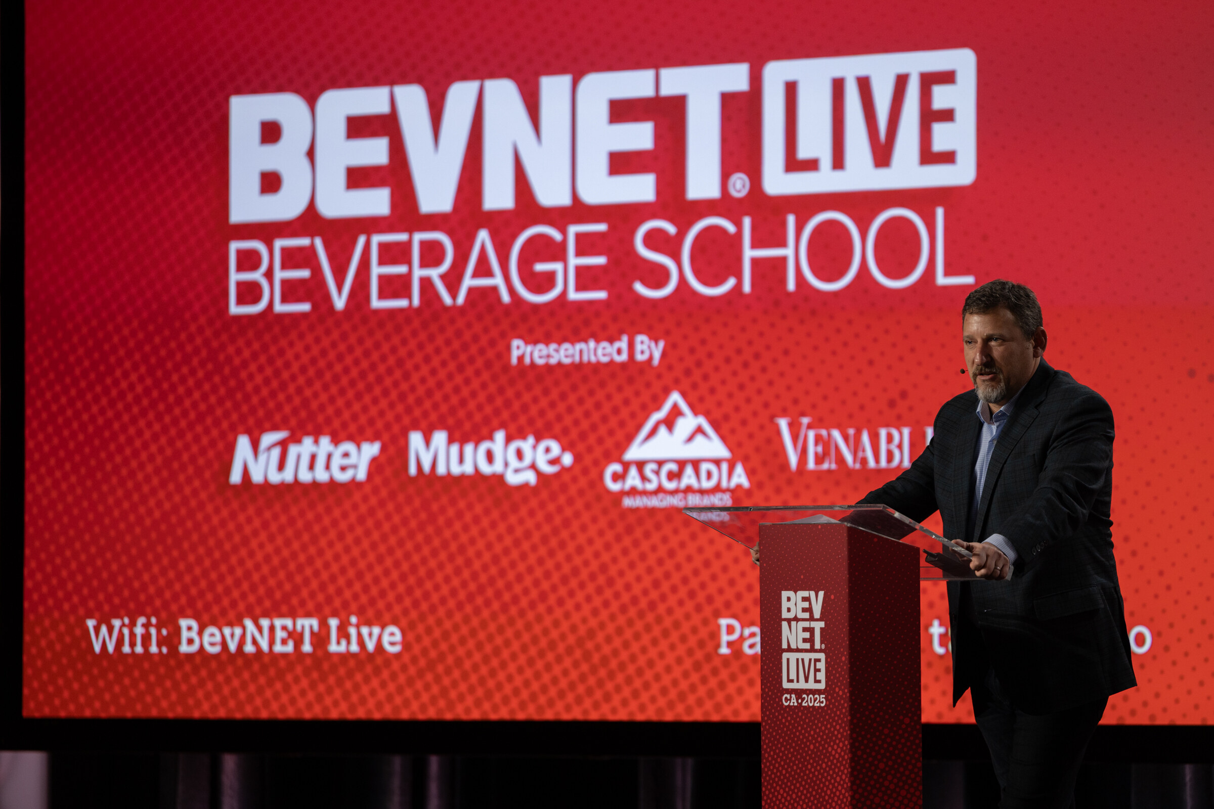 A man speaks at a podium beside a BevNET Live Beverage School screen.