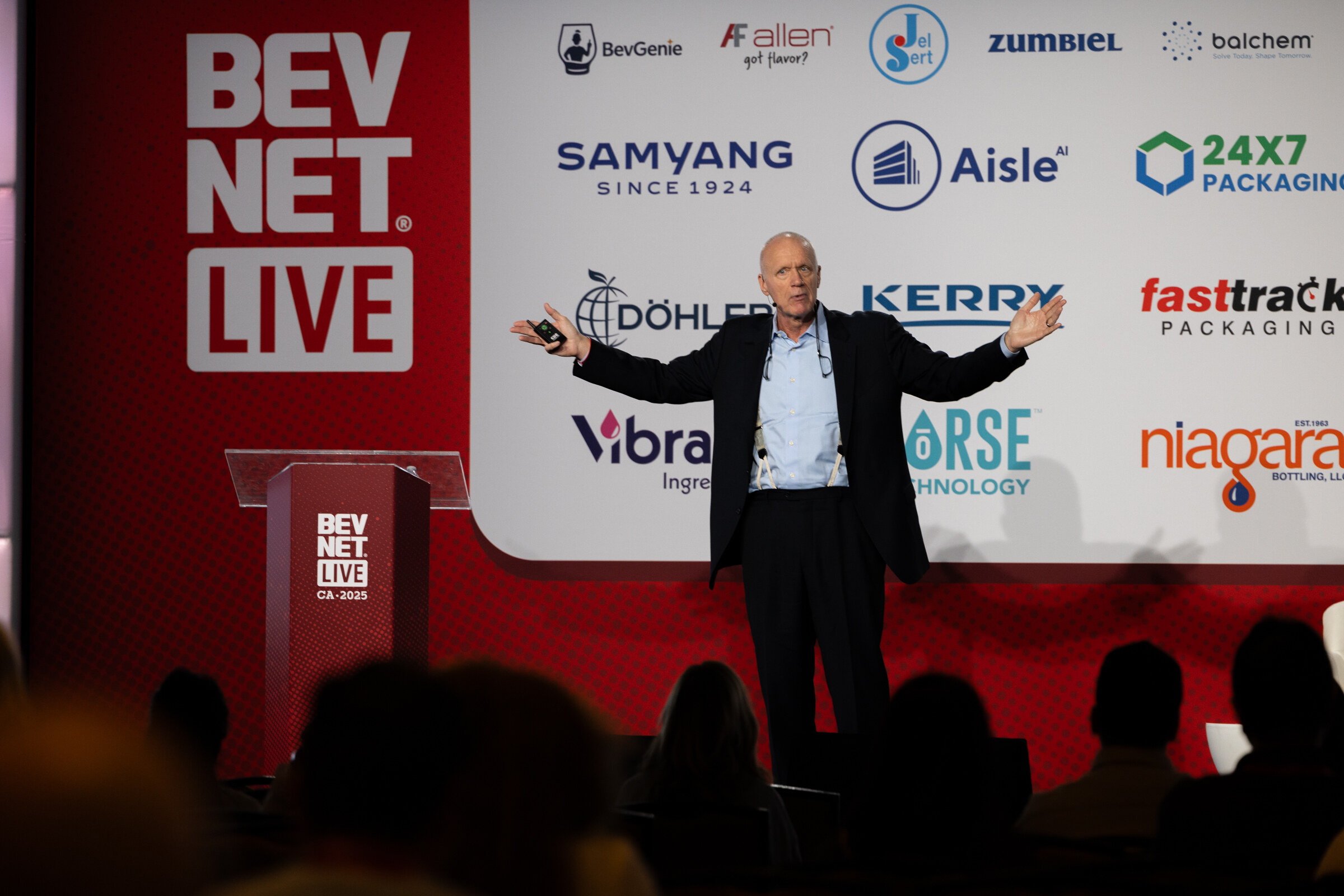 A bald man in a dark shirt spreads his arms while speaking on the Beverage School stage.