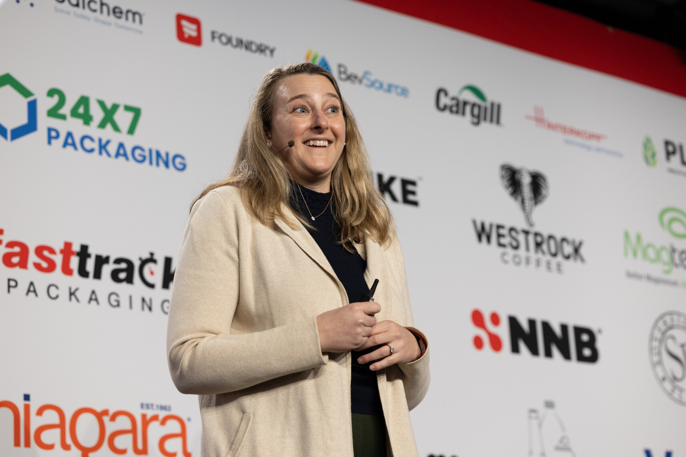 A woman in a cream blazer smiles while presenting in front of a sponsor wall.