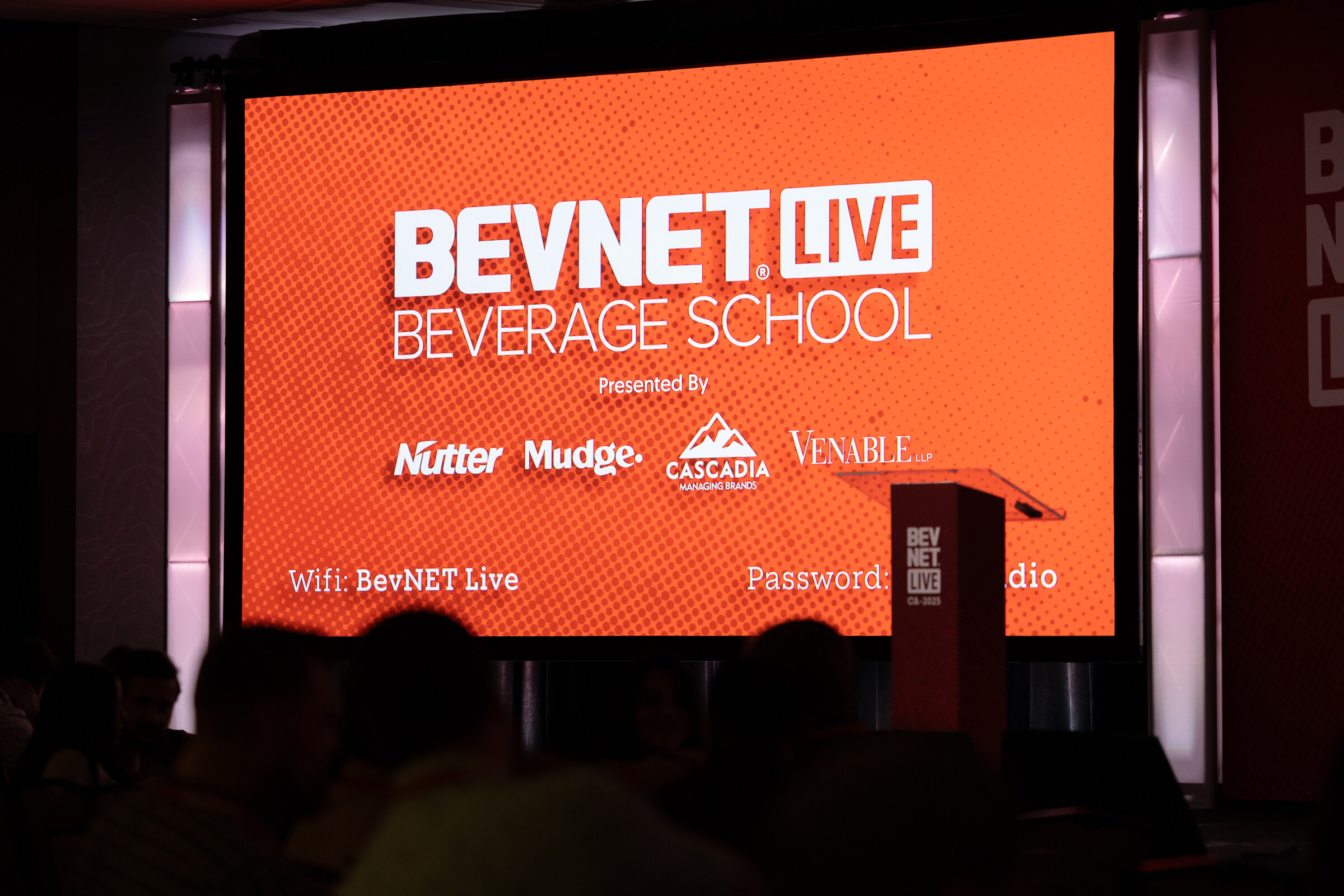 A red BevNET Live Beverage School slide glows above a podium on a darkened stage.