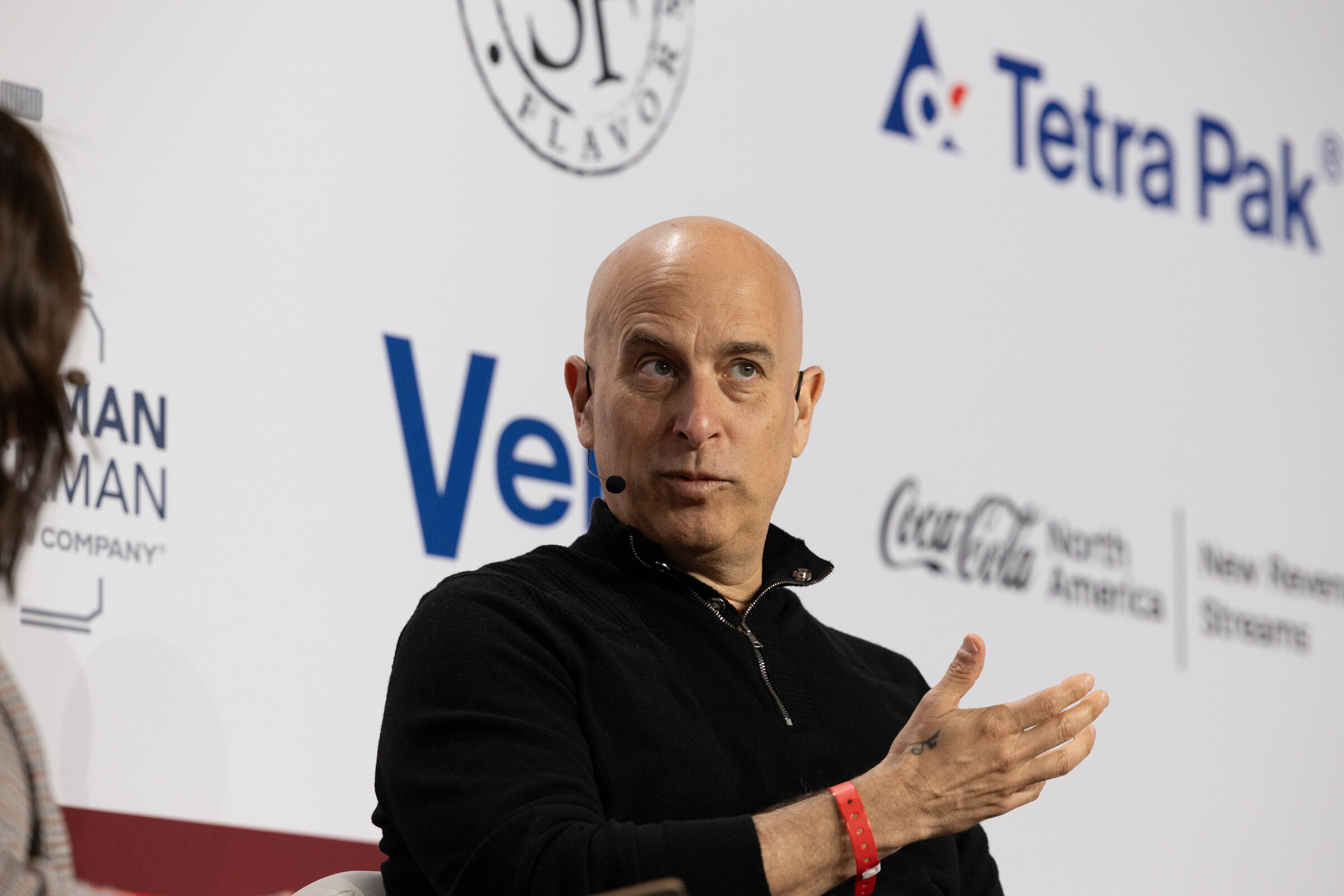 A bald man in a black sweater gestures while seated in front of a sponsor wall.