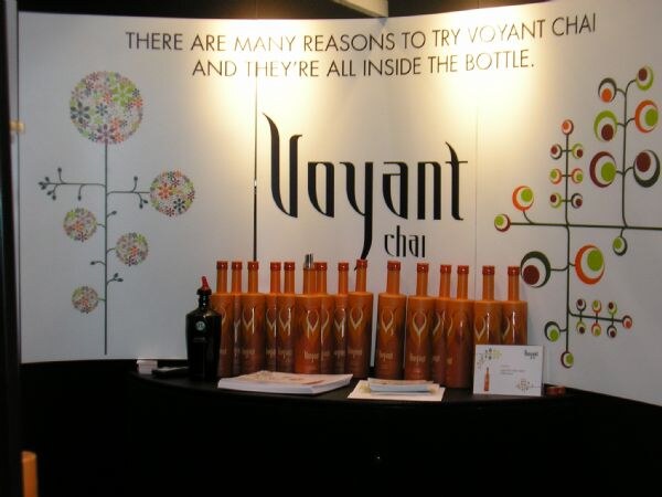 Voyant Chai booth with a curved white backdrop reading "There are many reasons to try Voyant Chai and they're all inside the bottle," and rows of orange bottles on a black-draped table.