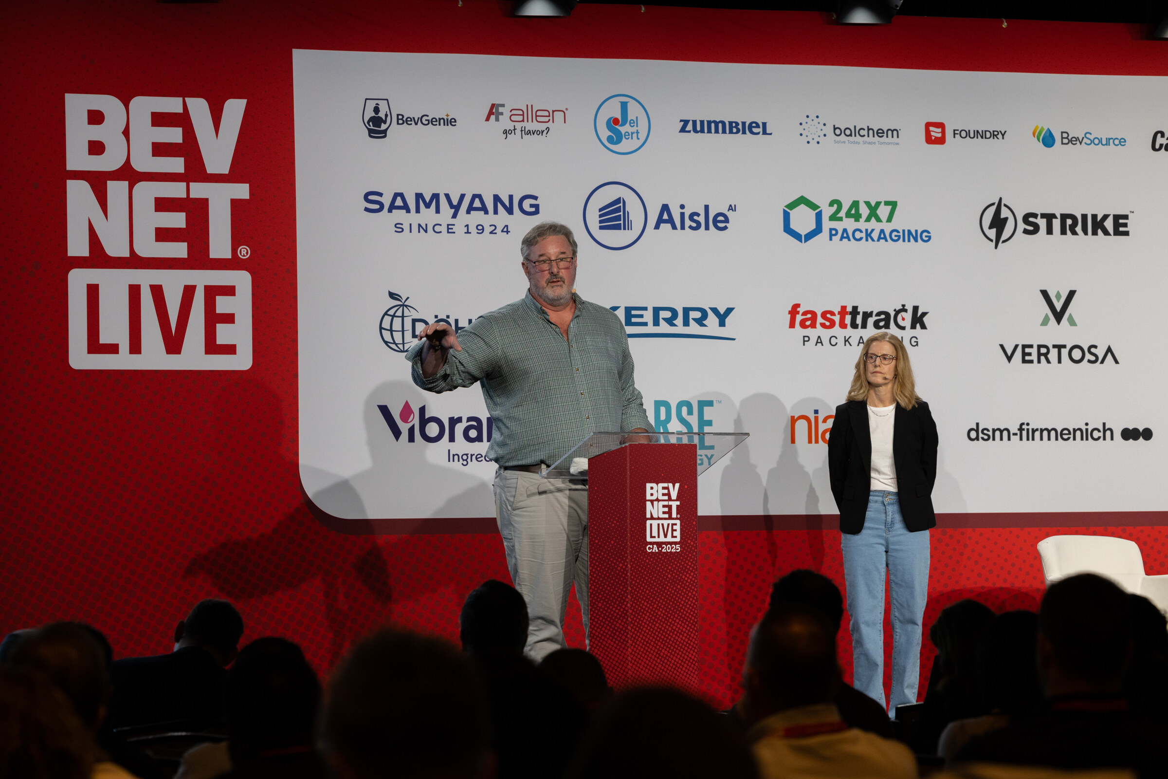 A man at the podium speaks while a woman stands beside him on stage.