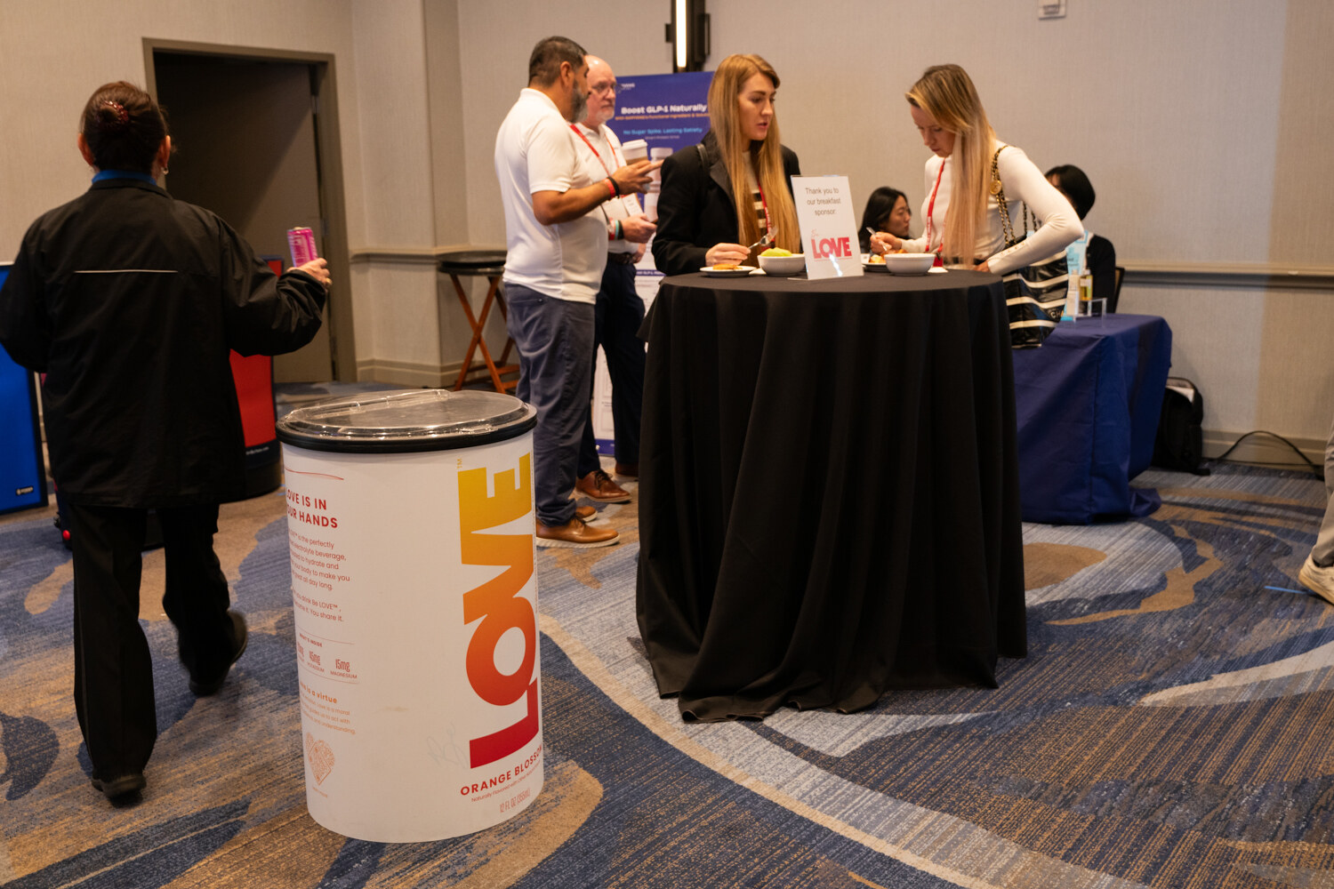 Attendees talk around a standing table beside a cylinder cooler printed with a Love logo.