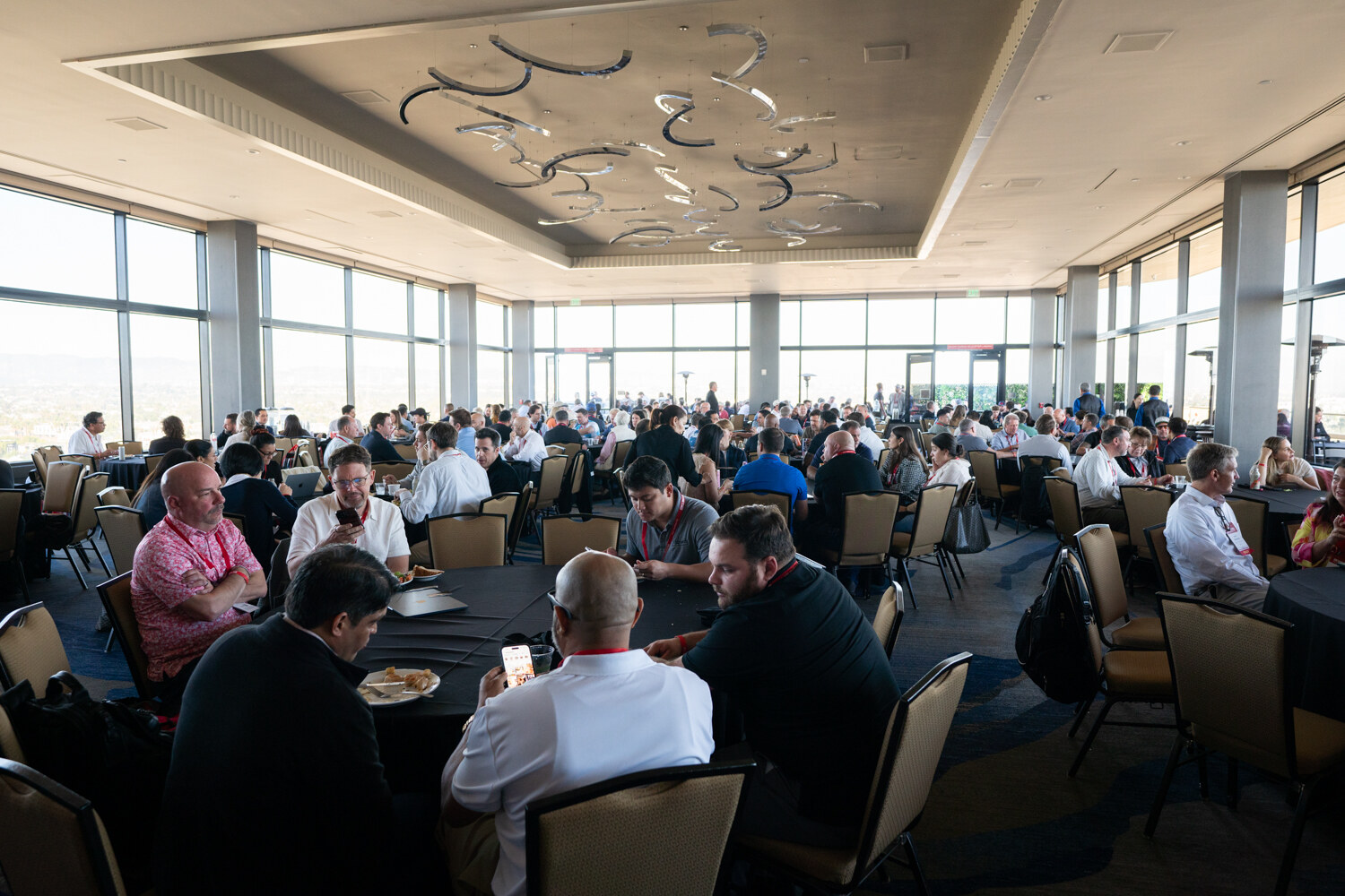 Attendees sit at round tables in a ballroom with large windows.