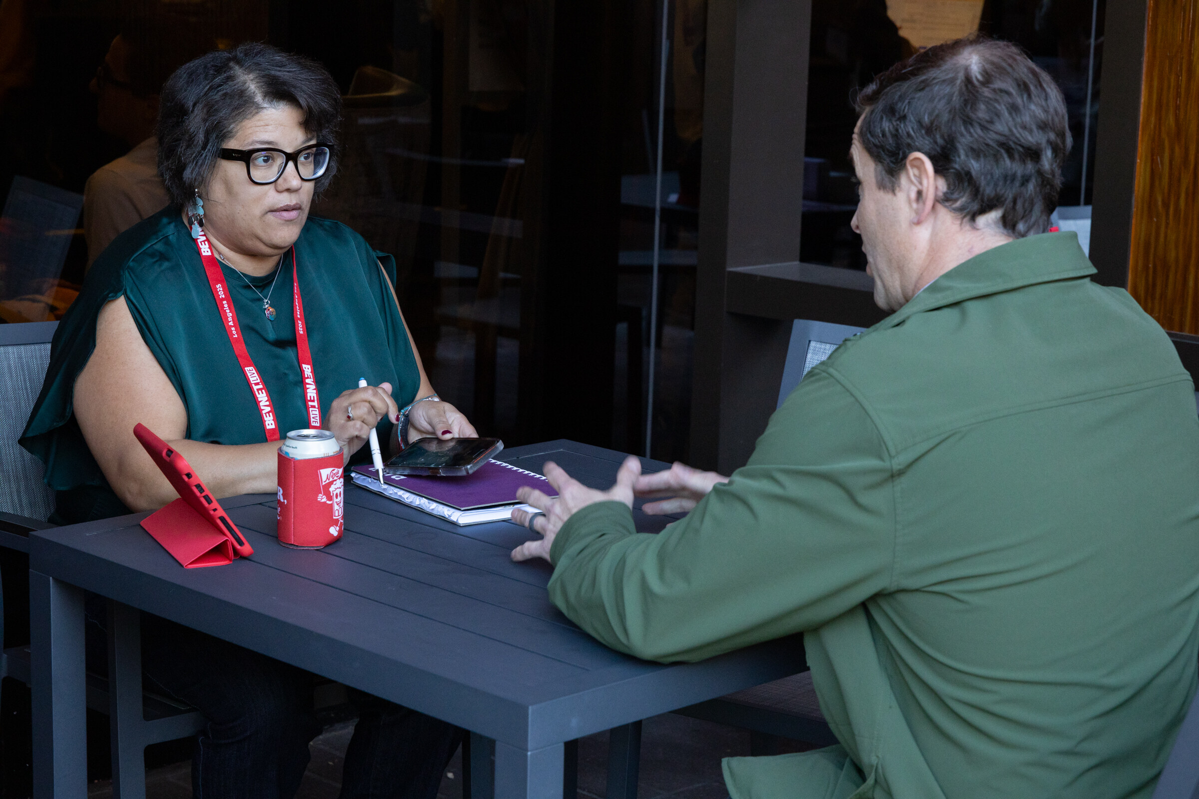 A woman in a green top and a man in a green jacket talk across a table.