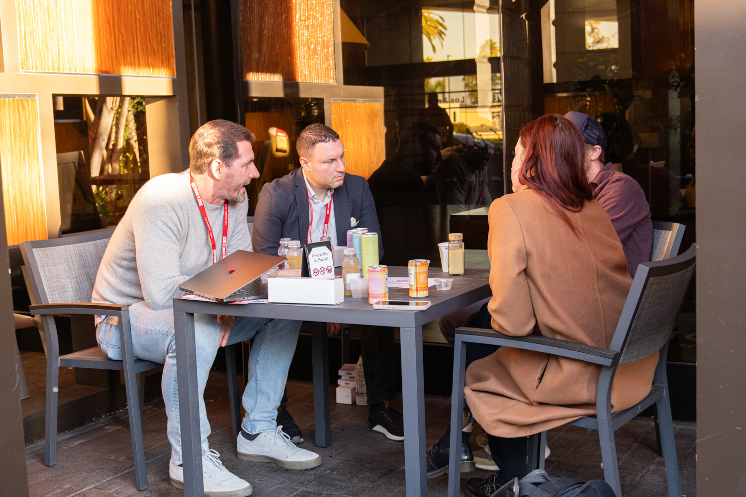 Four attendees talk over laptops and cans at a patio table.