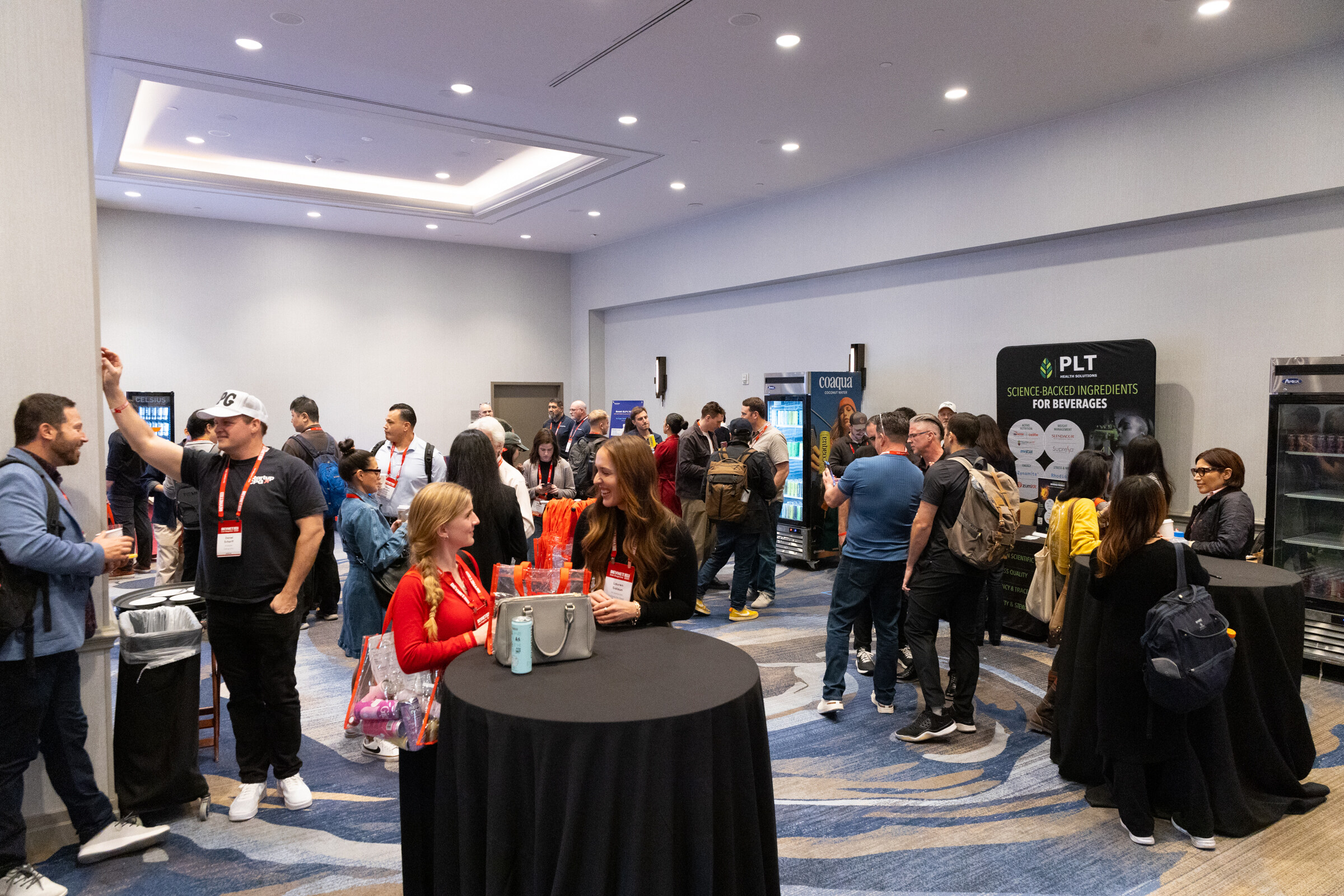 Attendees gather around cocktail tables and exhibitors in a meeting room.