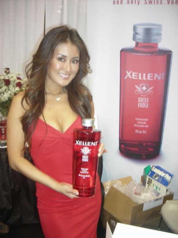 A smiling woman with long brown hair in a red dress holds a red bottle of Xellent Swiss Vodka beside a poster of the same bottle. Flowers and a cardboard box are nearby.