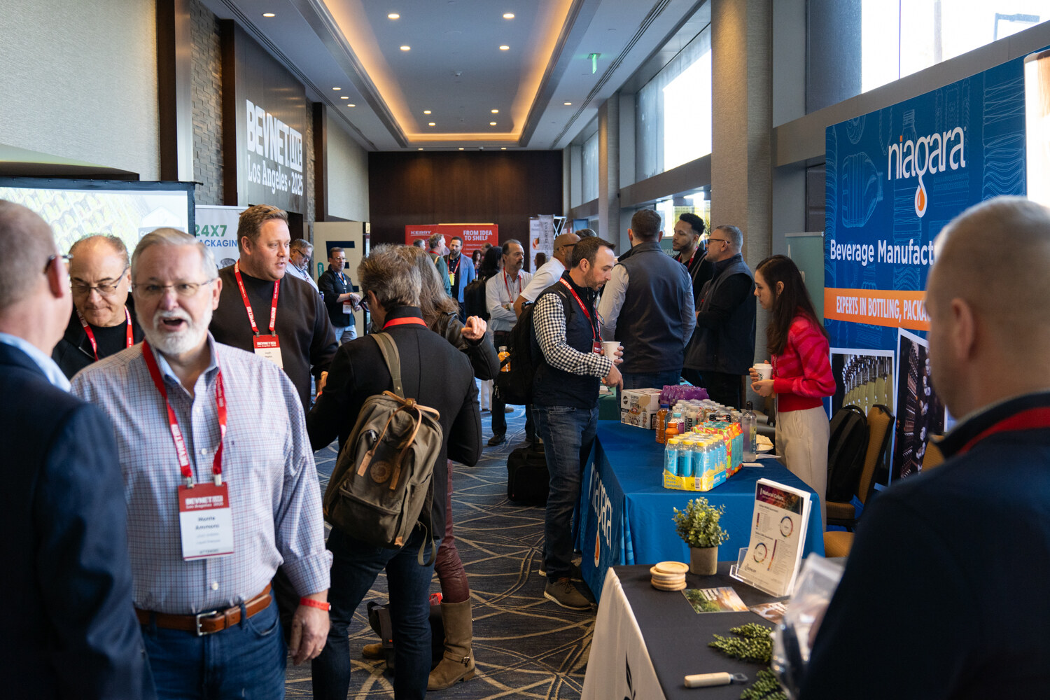 A man in a light shirt stands in a busy hallway lined with sponsor tables.