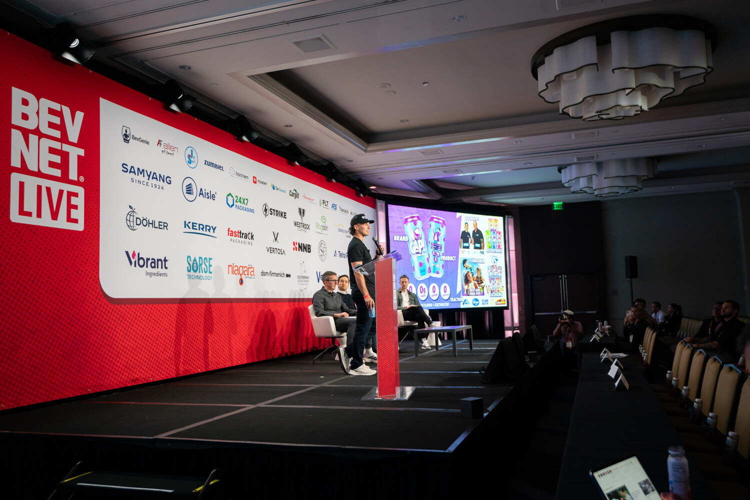 A wide view of the stage with a presenter, judges and a large screen.