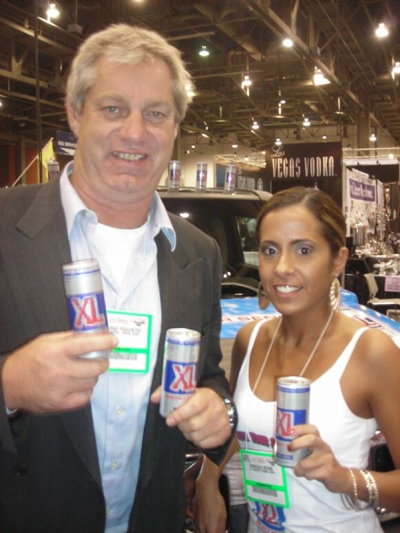 A man in a dark jacket and a woman in a white tank top each hold cans of XL energy drink, in front of a blue vehicle and a Las Vegas Vodka sign. Both wear exhibitor badges.