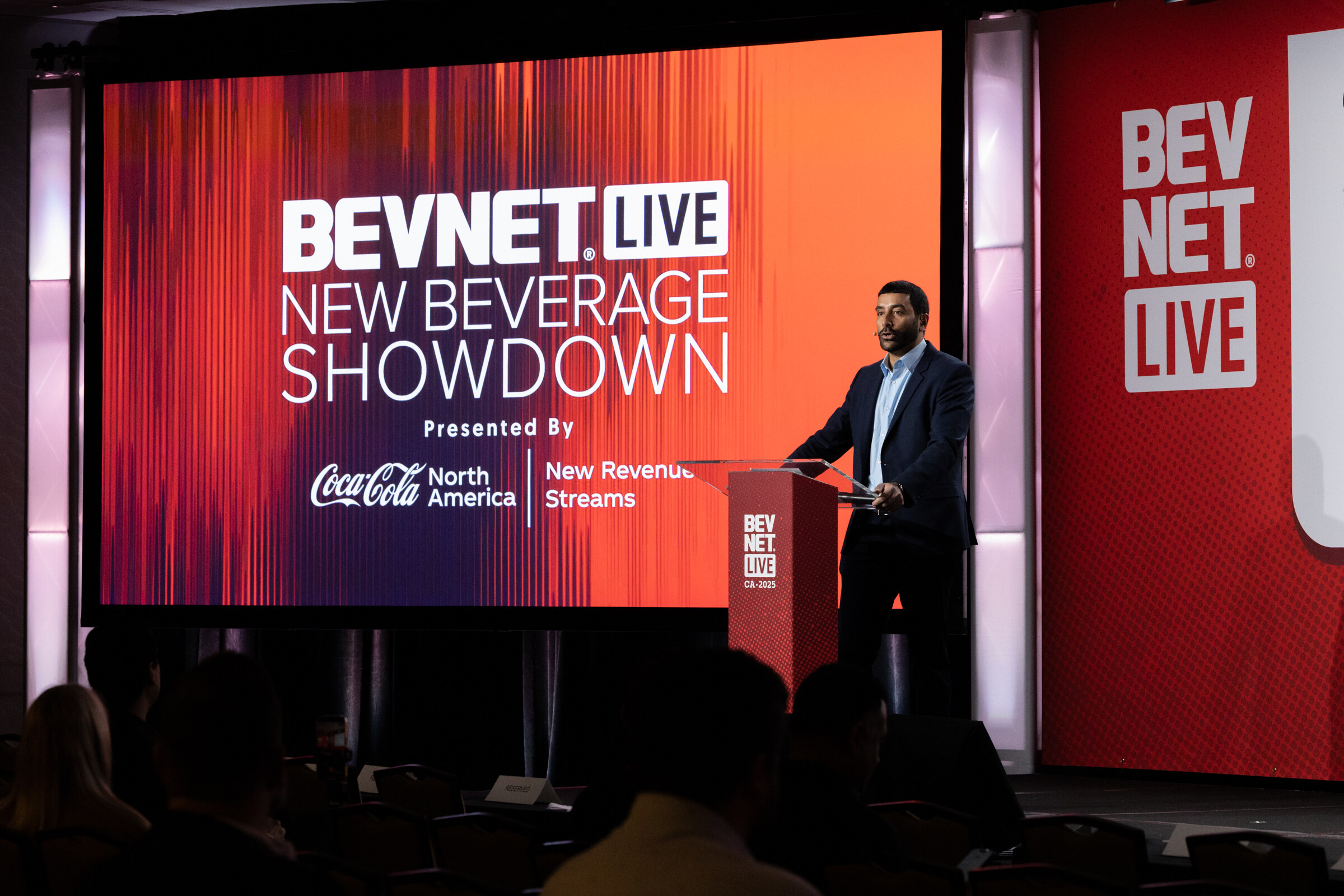 A man in a gray blazer stands at the podium in front of a New Beverage Showdown screen.