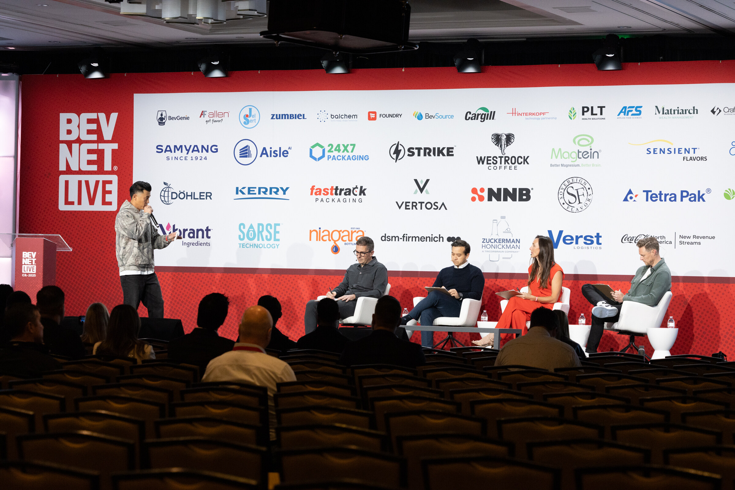 A presenter stands beside a panel of judges on a stage in a ballroom.