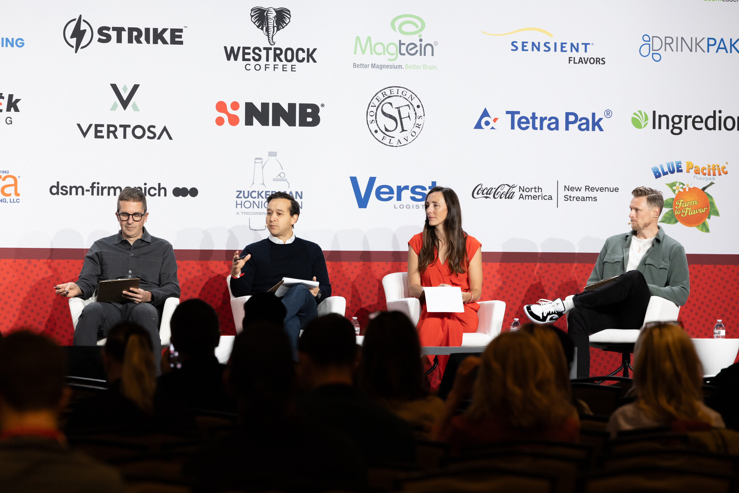 Four judges sit in white chairs on stage in front of a sponsor wall.