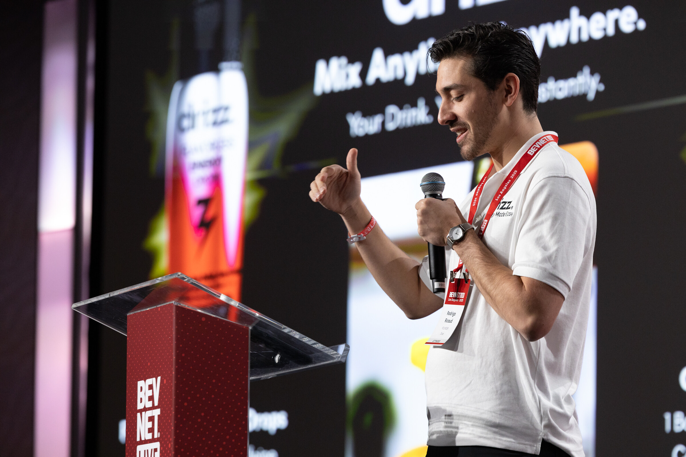 A man in a white shirt gives a thumbs-up at a lectern in front of a drink slide.