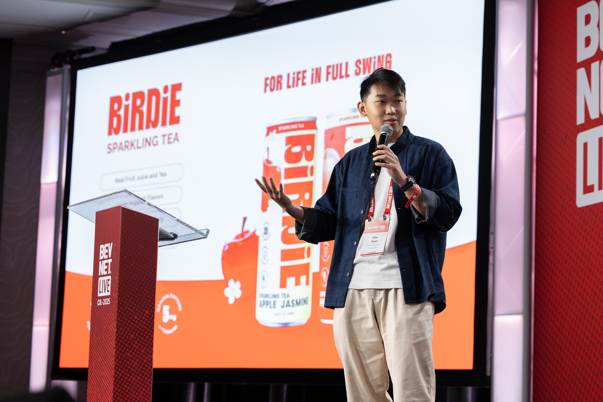 A man in a navy jacket speaks at a lectern in front of a Birdie slide.