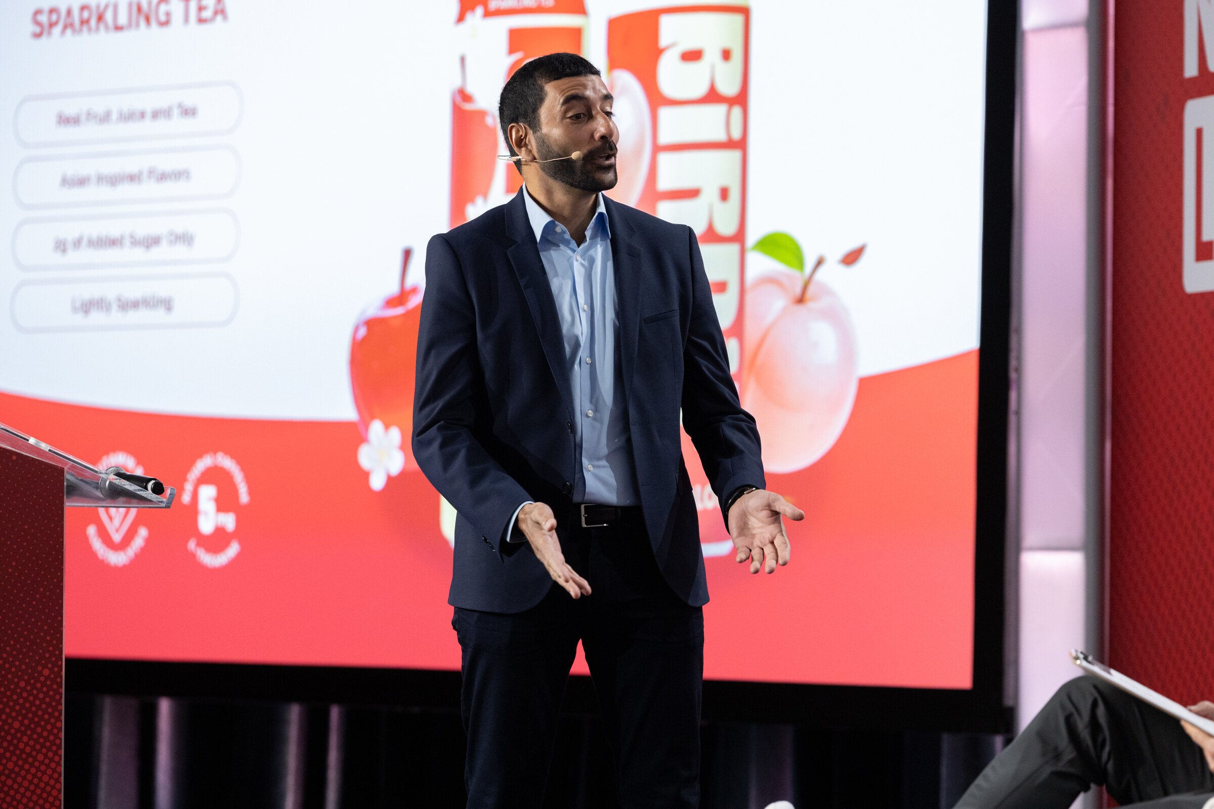 A man in a dark suit gestures in front of a sparkling tea slide.