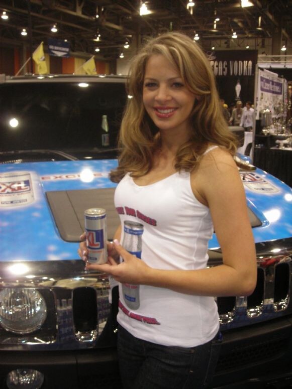 A smiling woman in a white tank top holds a can of XL energy drink in front of a vehicle with a blue XL-branded hood. A Las Vegas Vodka sign is in the background.