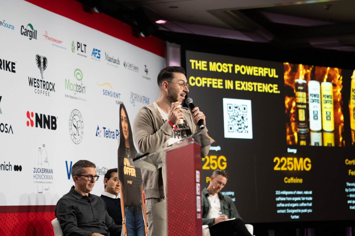 A presenter stands at the lectern in front of a Power Coffee slide while judges sit nearby.