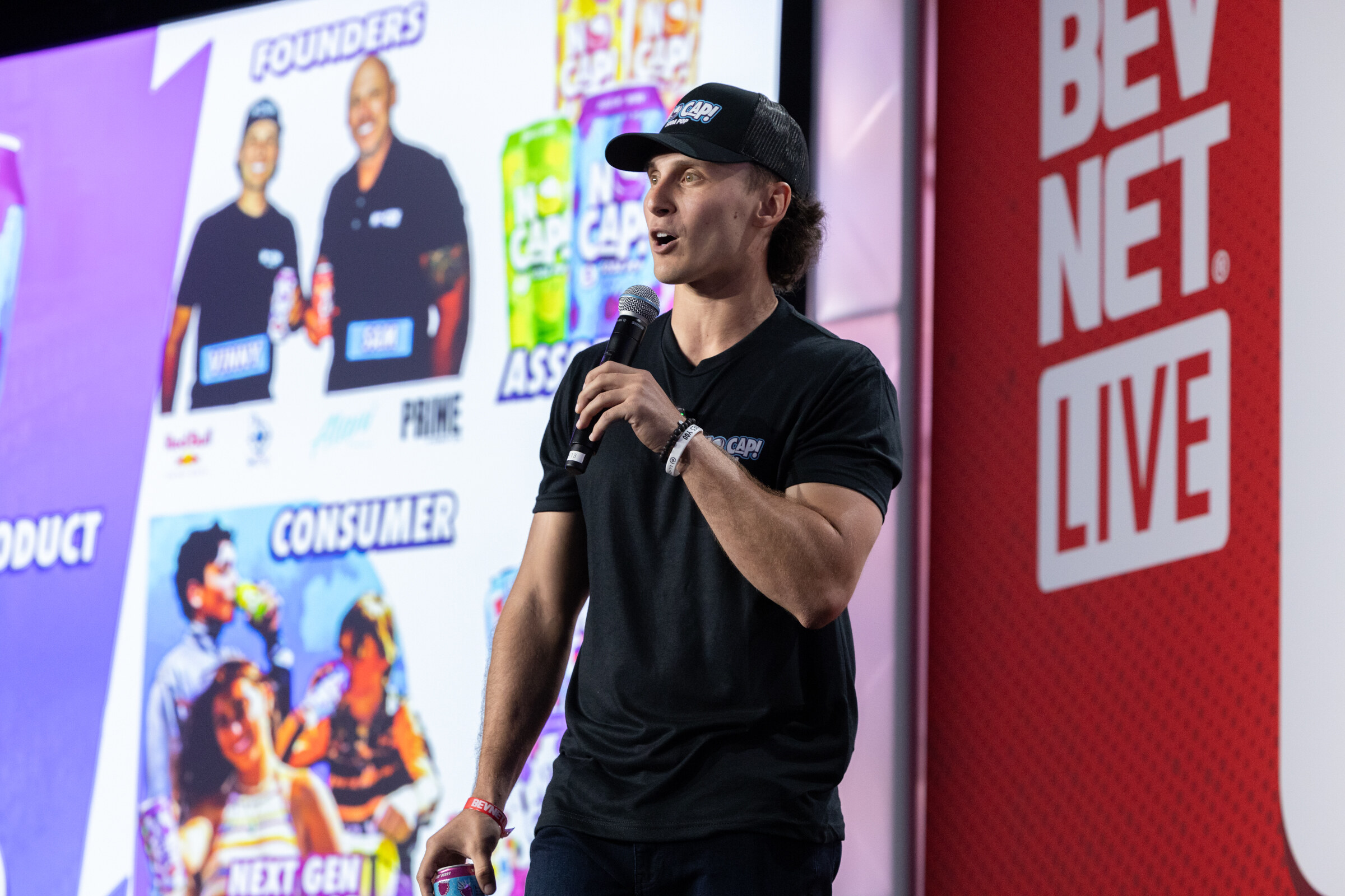 A man in a black shirt and cap speaks in front of a slide of founders.