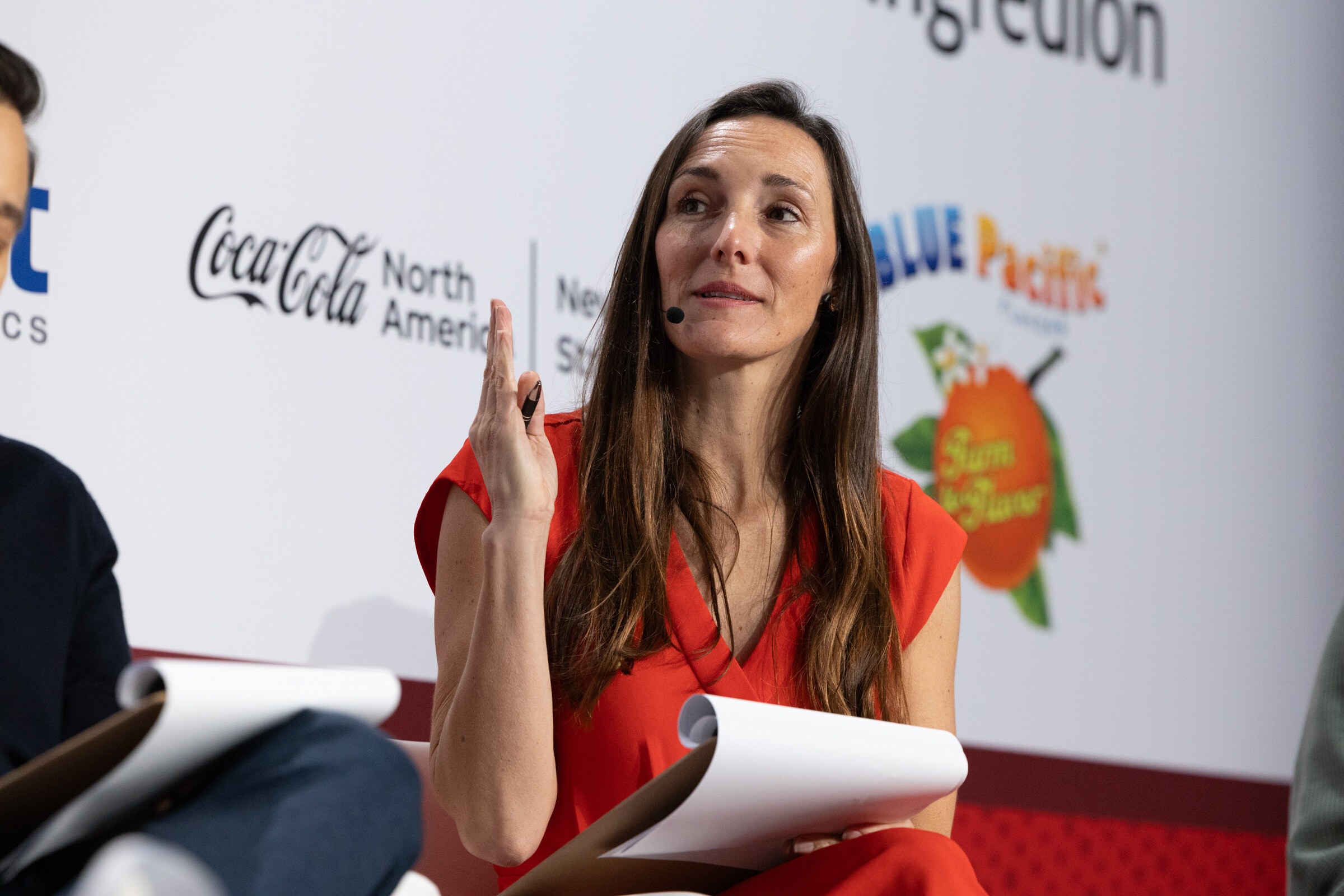 A woman in a red dress gestures while speaking into a headset mic.