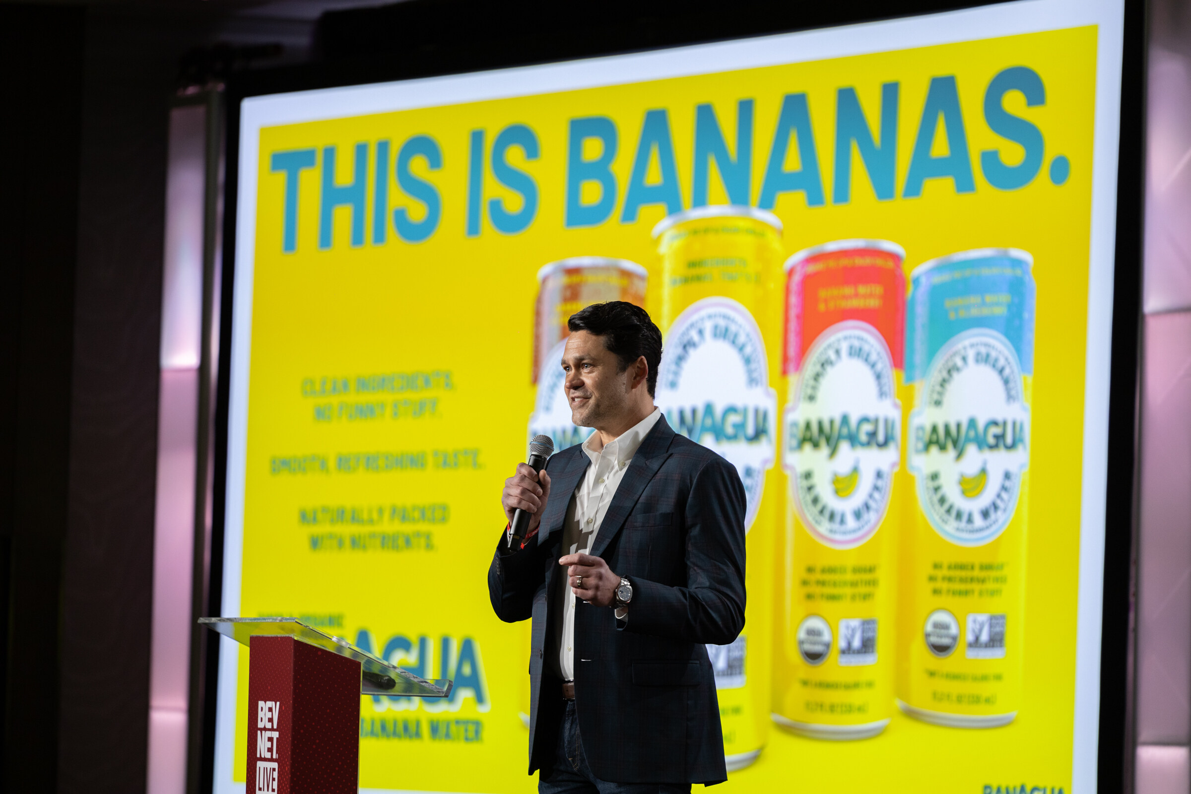 A man in a dark suit speaks in front of a bright yellow Banagua slide.