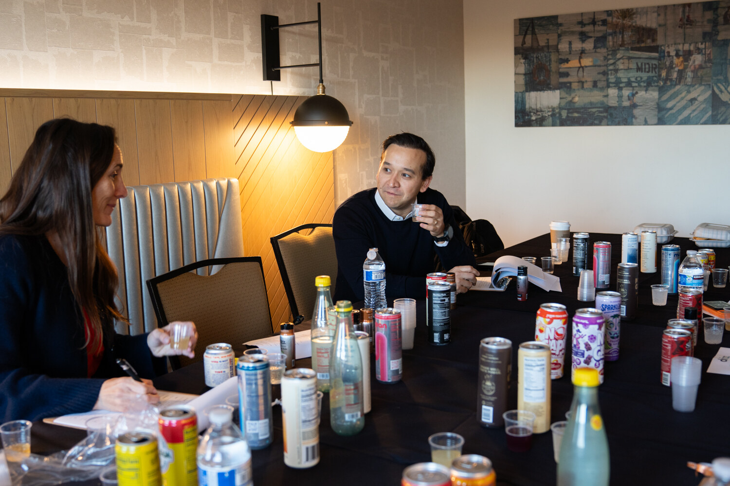 A man and a woman at a long table with many cans and cups taste products.