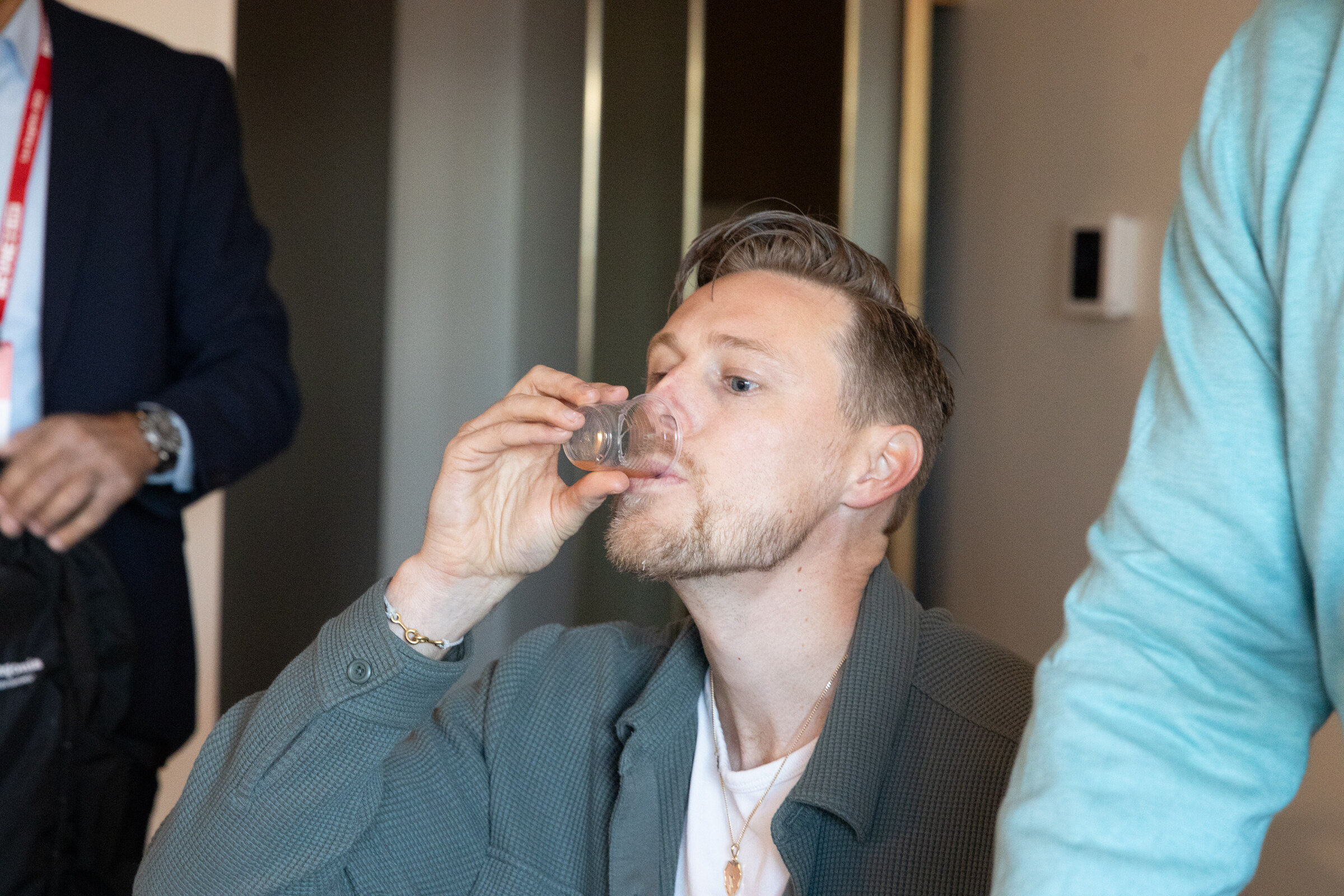 A man in a green jacket drinks from a small cup.