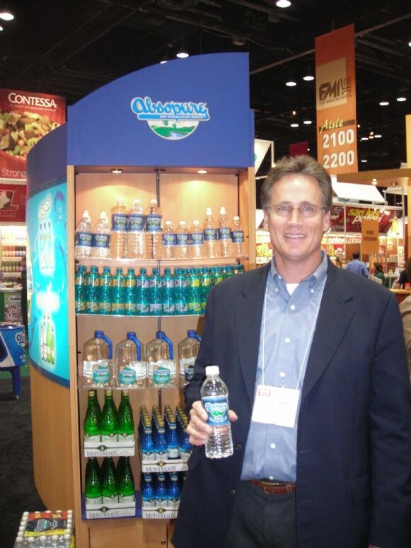 A smiling man with glasses and a blazer holds a bottle of Absopure water beside a display shelf of Absopure and Montreaux bottles. A Contessa banner is at left and an FMI aisle sign behind.