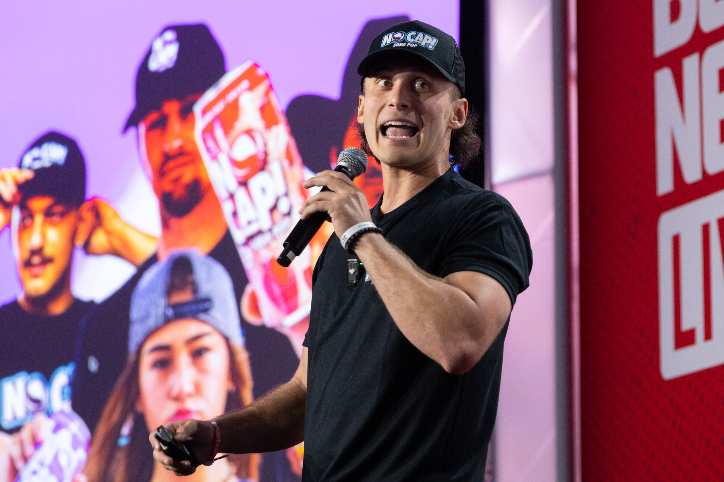 A man in a black cap and shirt speaks in front of a No Cap slide.