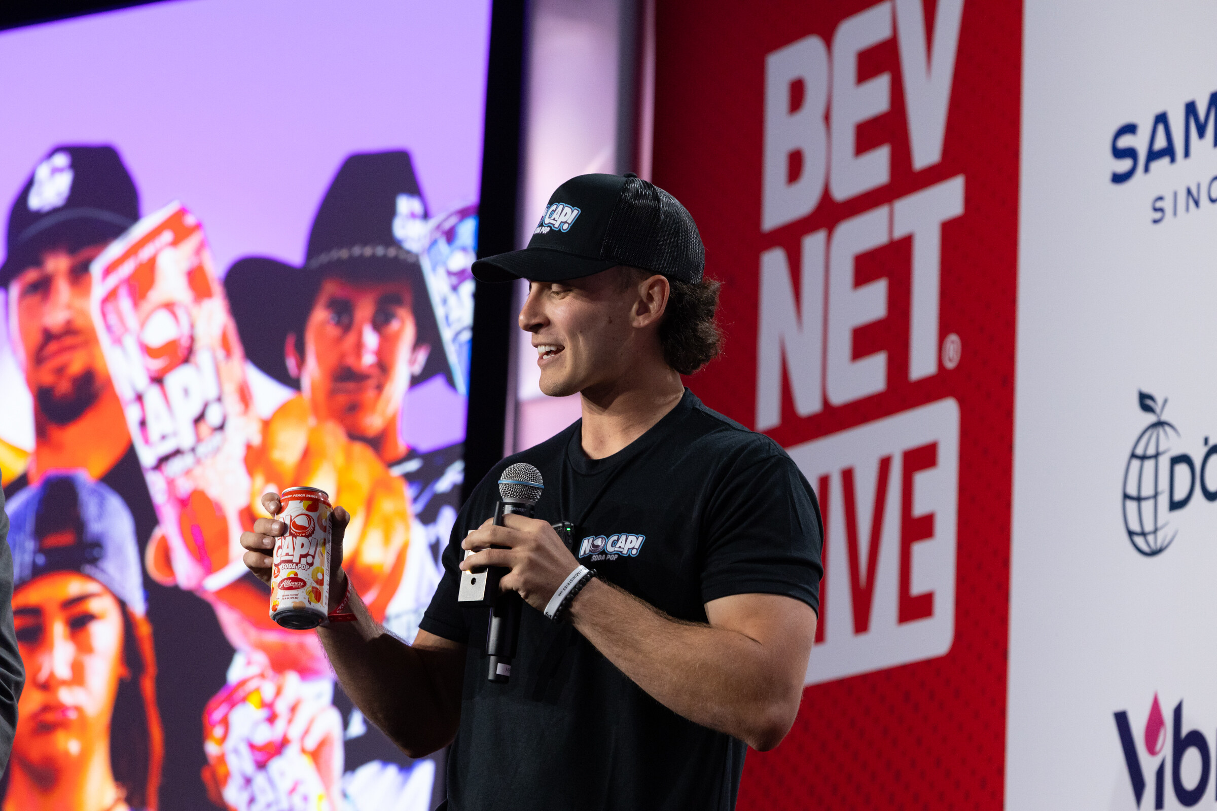 A man in a black cap holds a can while presenting.