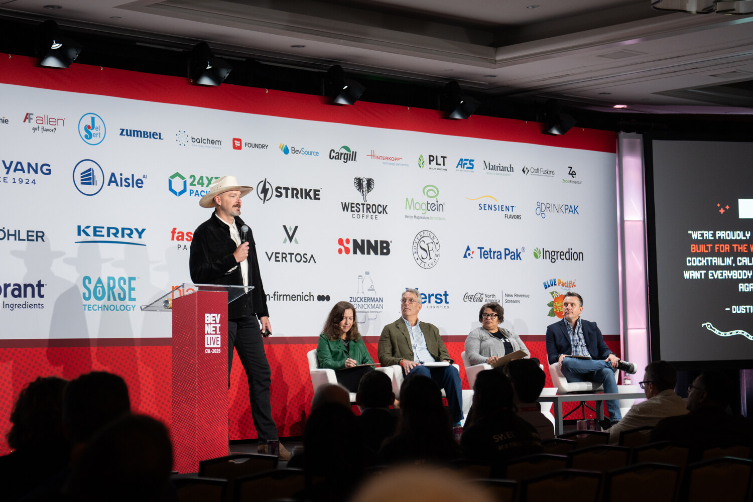 A wide shot shows a presenter at the lectern and judges seated on stage.