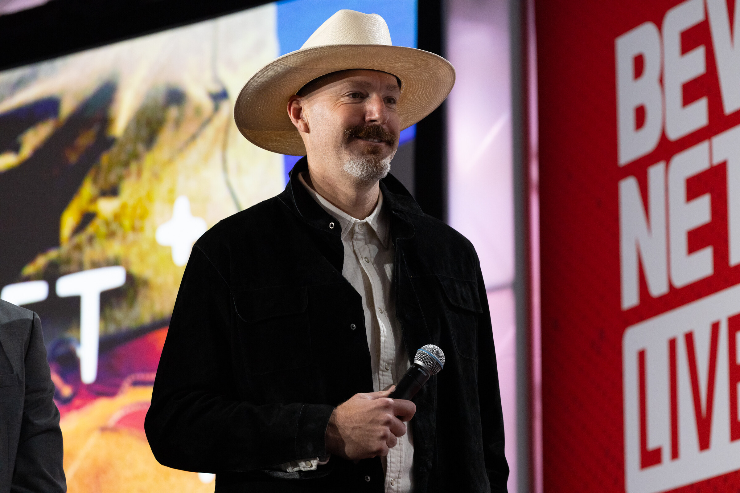 A man in a cowboy hat holds a microphone on stage.