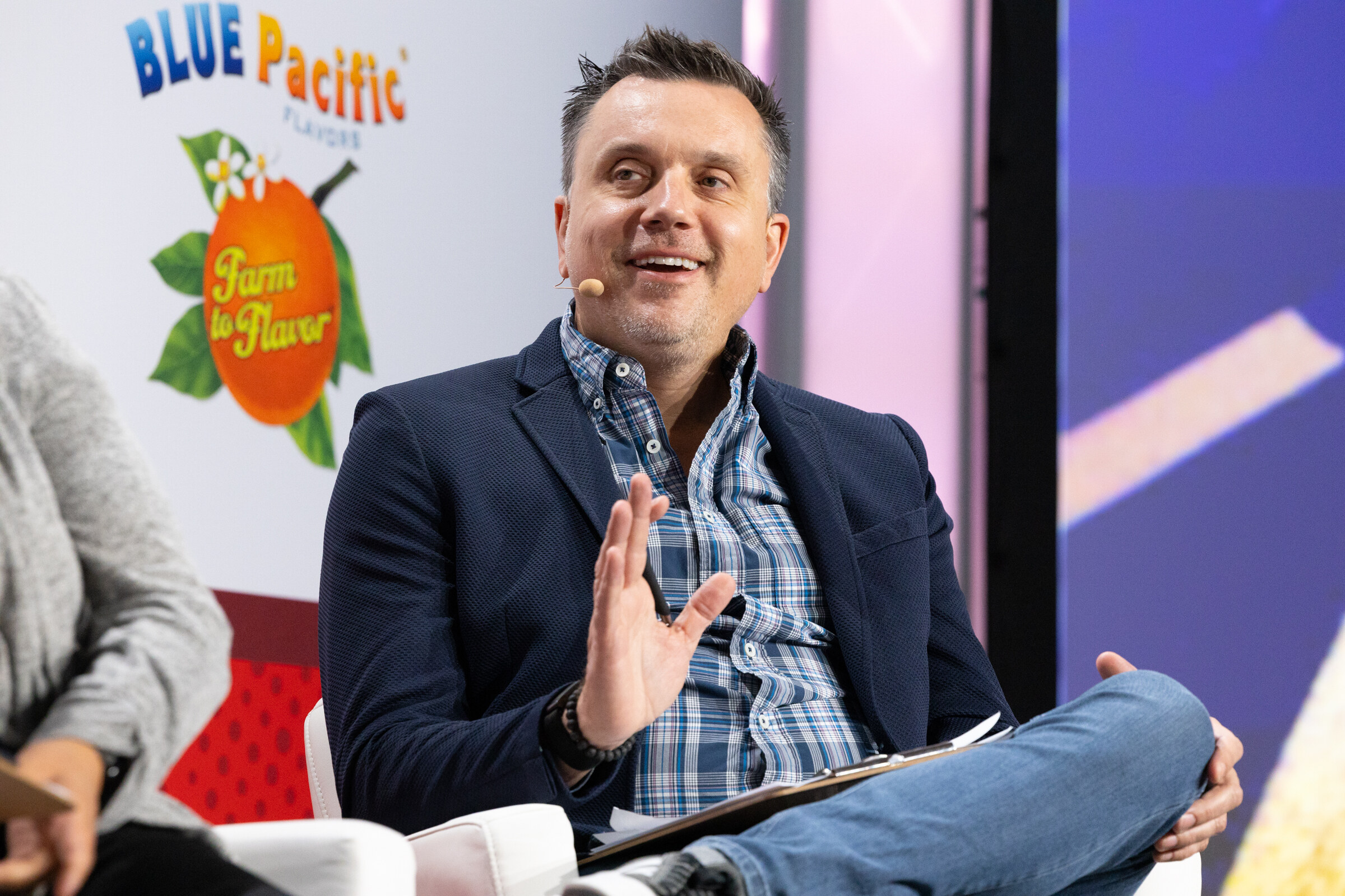 A man in a navy blazer and plaid shirt gestures while seated.