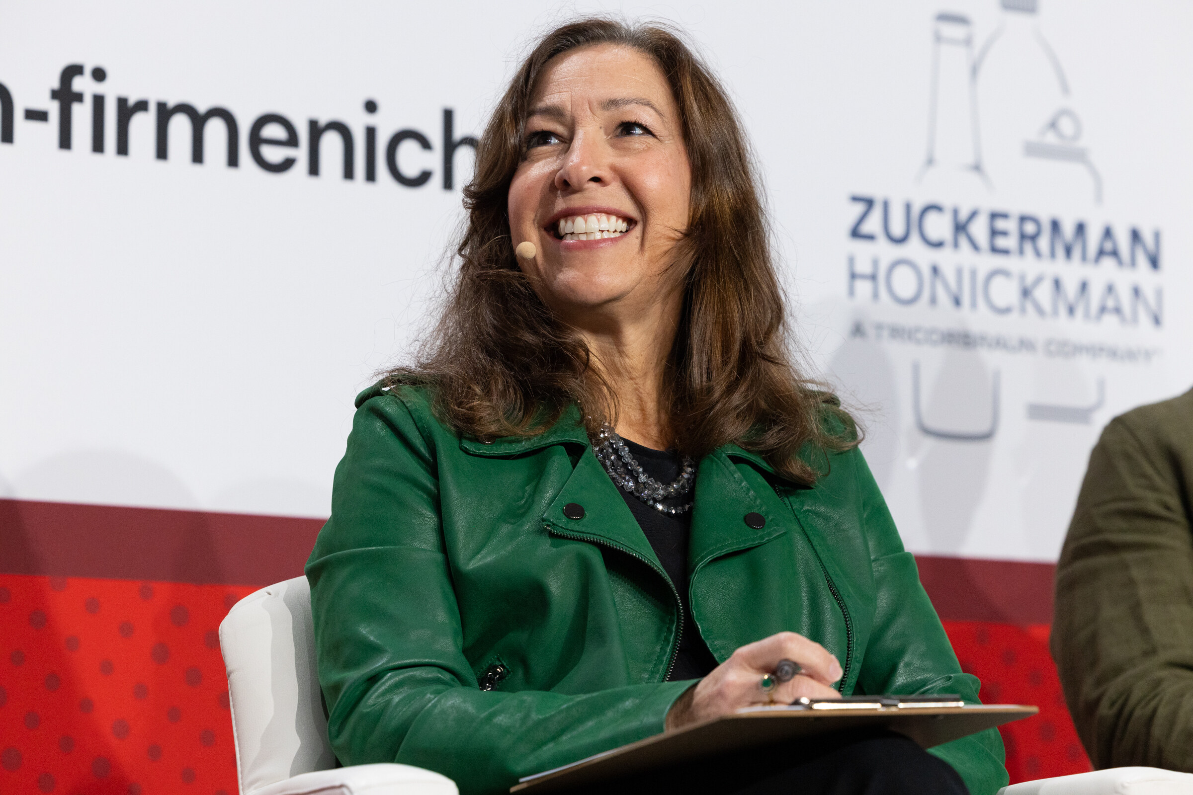 A woman in a green jacket smiles while seated with a clipboard.