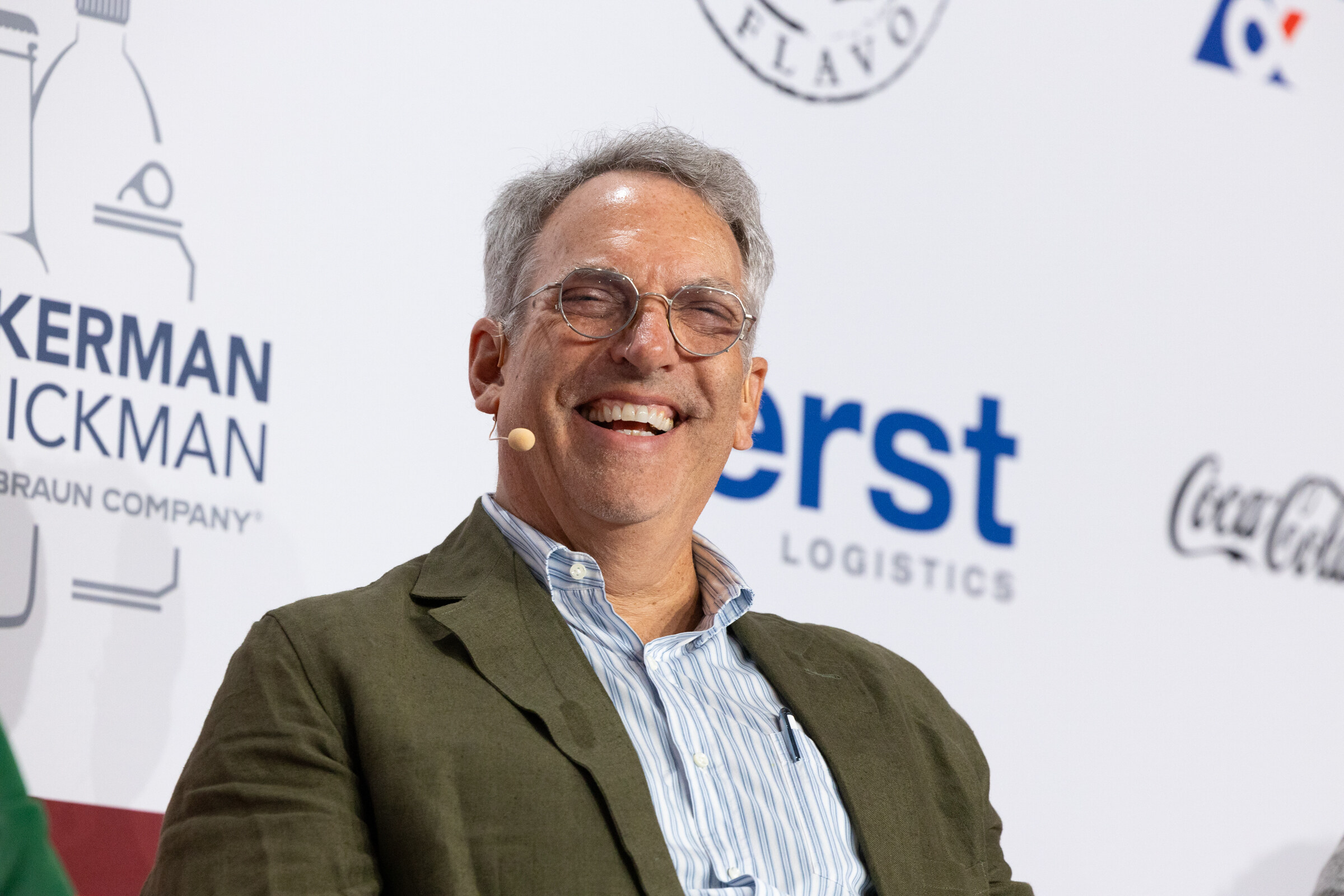 A gray-haired man in an olive jacket laughs on stage.