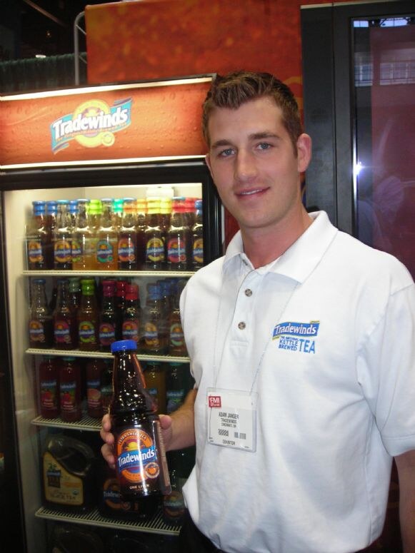 Adam Janser of Tradewinds, in a white Tradewinds polo, holds a bottle of Tradewinds tea beside a lit refrigerated display case filled with bottled teas.