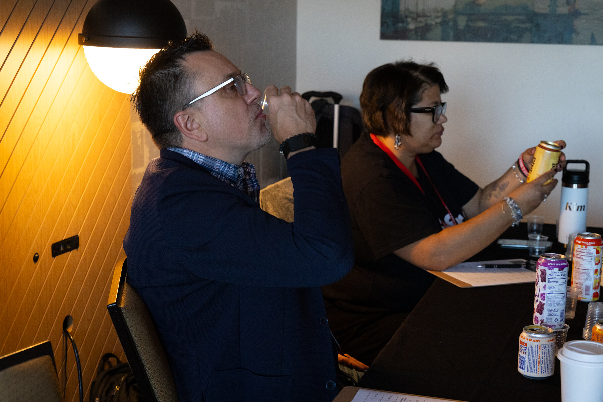 A man drinks from a can while a woman examines another can at a conference table.