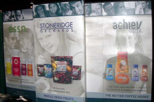Three tall backlit panels for essn, Stoneridge Orchards whole dried fruits and achiev ONE coffee boost, each showing product packages and photos of smiling people.