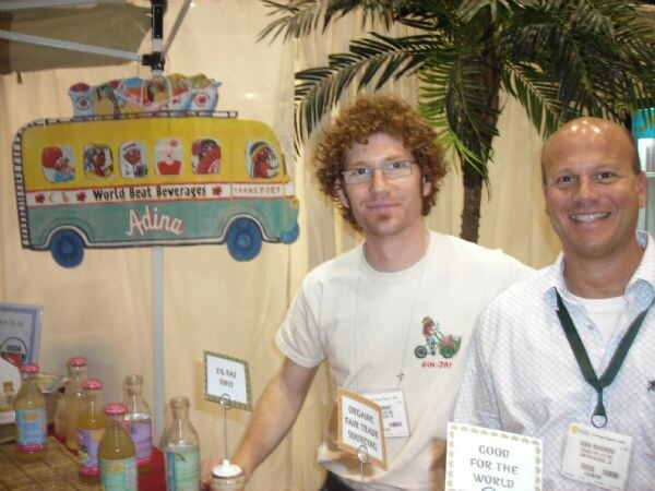 Two smiling men wearing badges stand at a booth with a painted World Beat Beverages Adina bus sign, a palm tree, and bottles on a table. Signs read "Good for the world."