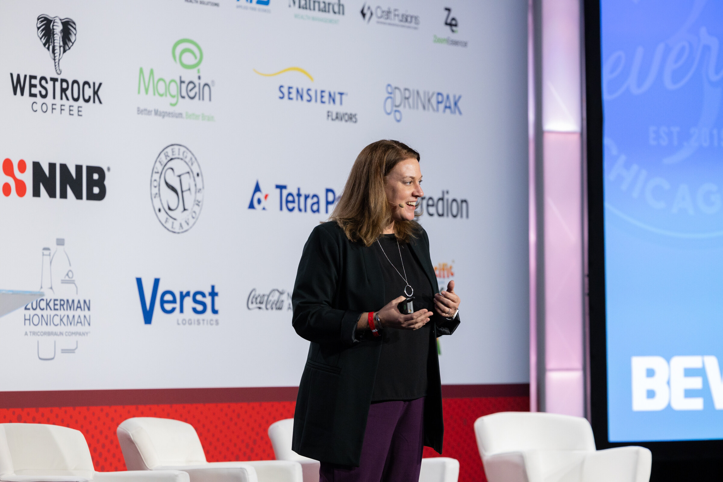 A woman in black presents on a large stage in front of a sponsor wall.