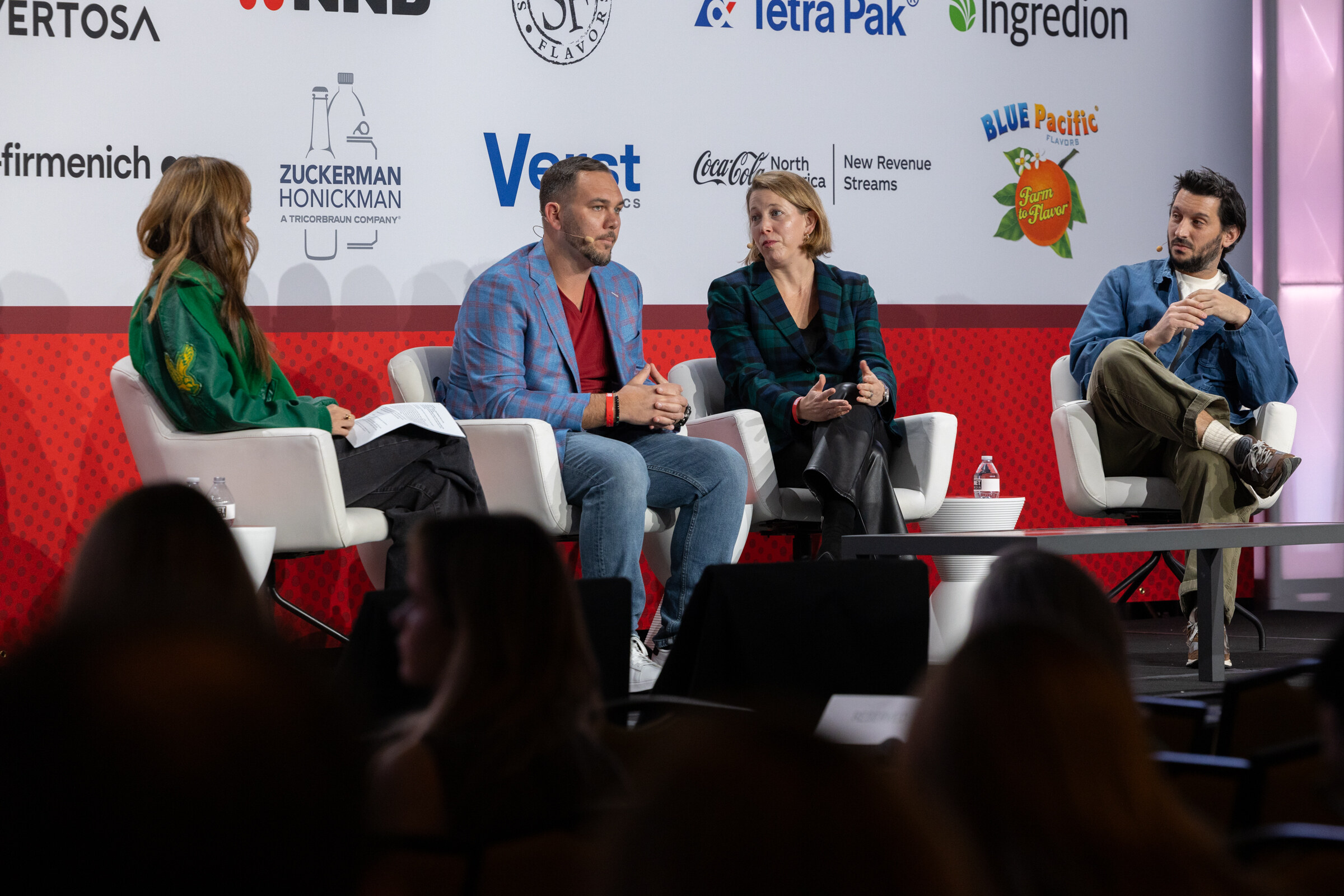 Four panelists sit in white chairs on stage in front of a sponsor backdrop.